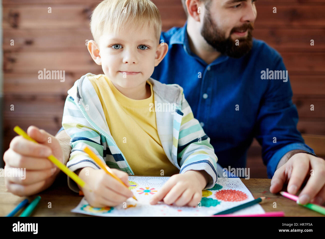 Niedlichen kleinen blonden Jungen mit tief graue Augen sitzen auf Schoß des Vaters bärtigen, Blick in die Kamera und halten Bleistift in der hand zum Färben von pict Stockfoto