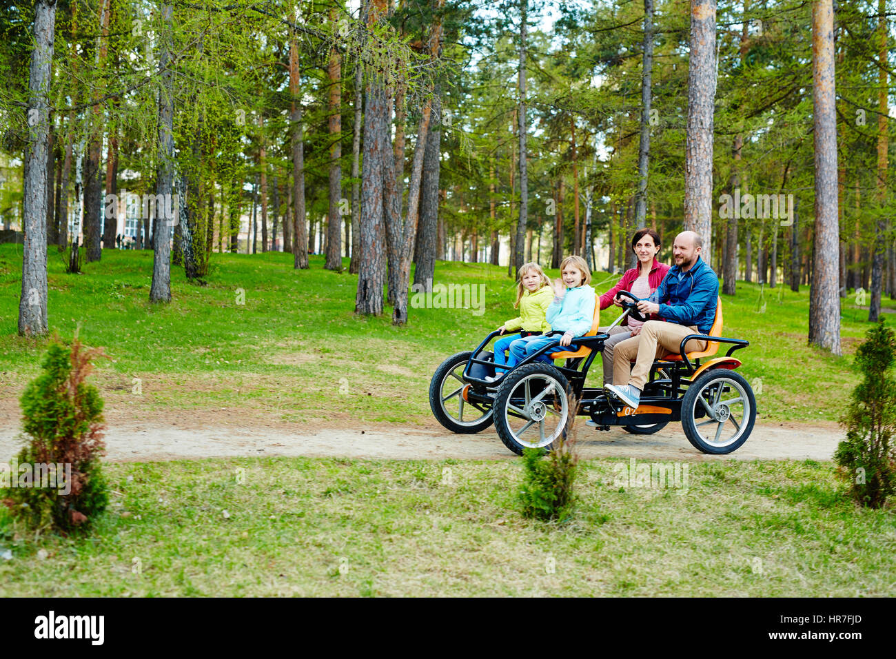 Fröhliche Familie von vier Reiten orange vierrädrige Tretauto genießen Frühling im Park mit Pinienwäldern Stockfoto