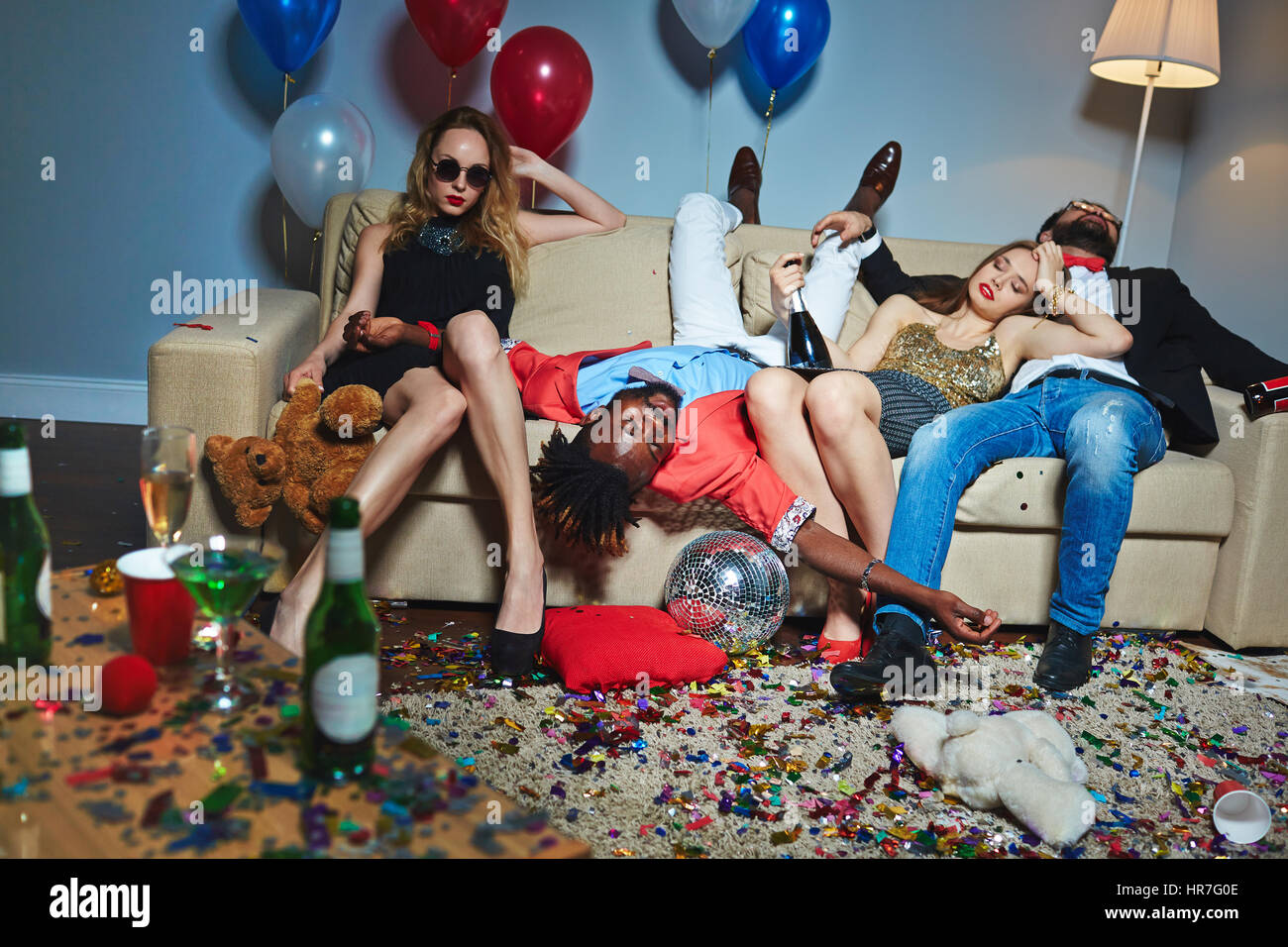 Unordentlicher Raum nach wilde Hausparty, tragen drei beschwipst stilvolle Freunde entspannend auf Couch während der blonden Frau mit Teddy posiert für Fotografie Stockfoto