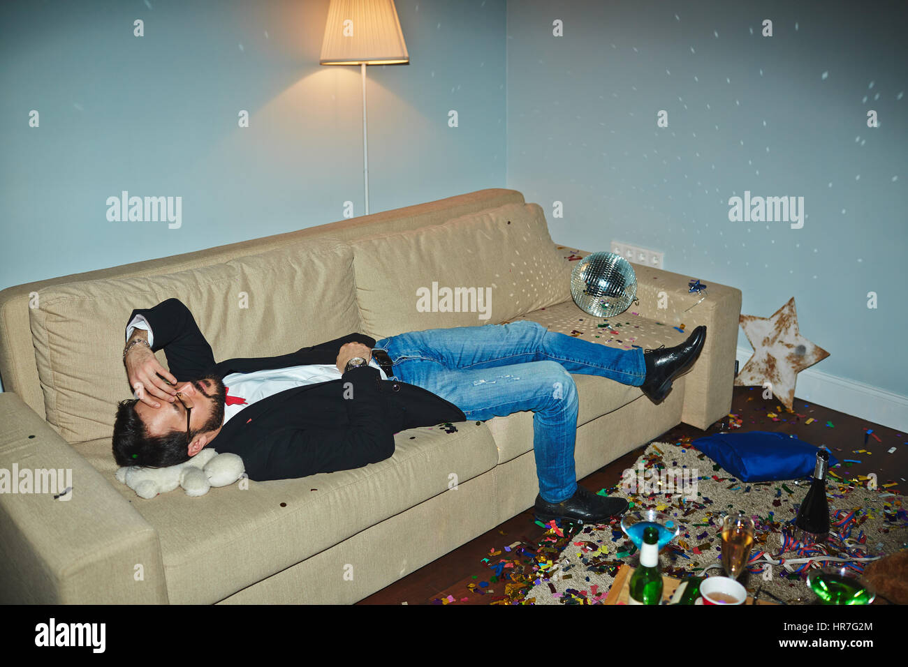 Asiatischer Mann in Jeans und Jacke mit Nickerchen an chaotisch Wohnzimmer nach wilde Silvester-party Stockfoto