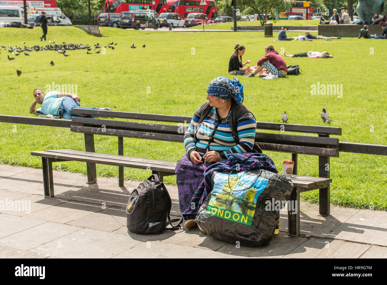 Eine östliche europäische Frau ein Kopftuch sitzen auf einer Bank in einem Park in London mit ihrem Gepäck. Stockfoto