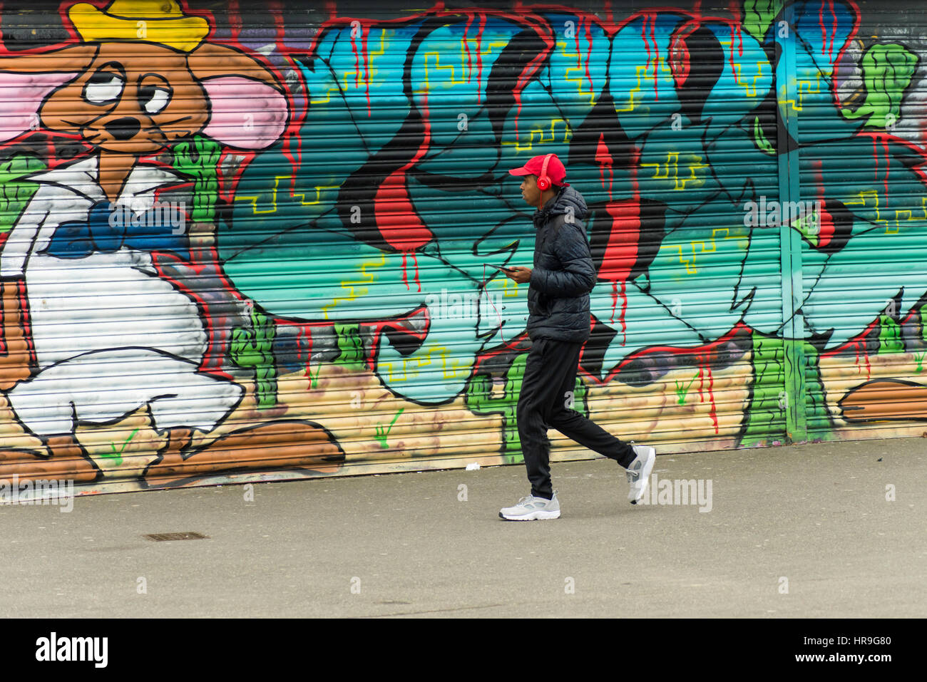 Eine junge schwarze Männer mit Kopfhörern zu Fuß vorbei an bunten Graffiti auf der Vorderseite eines stillgelegten Shop direkt an der Küste in Ramsgate, Kent, Großbritannien. Stockfoto