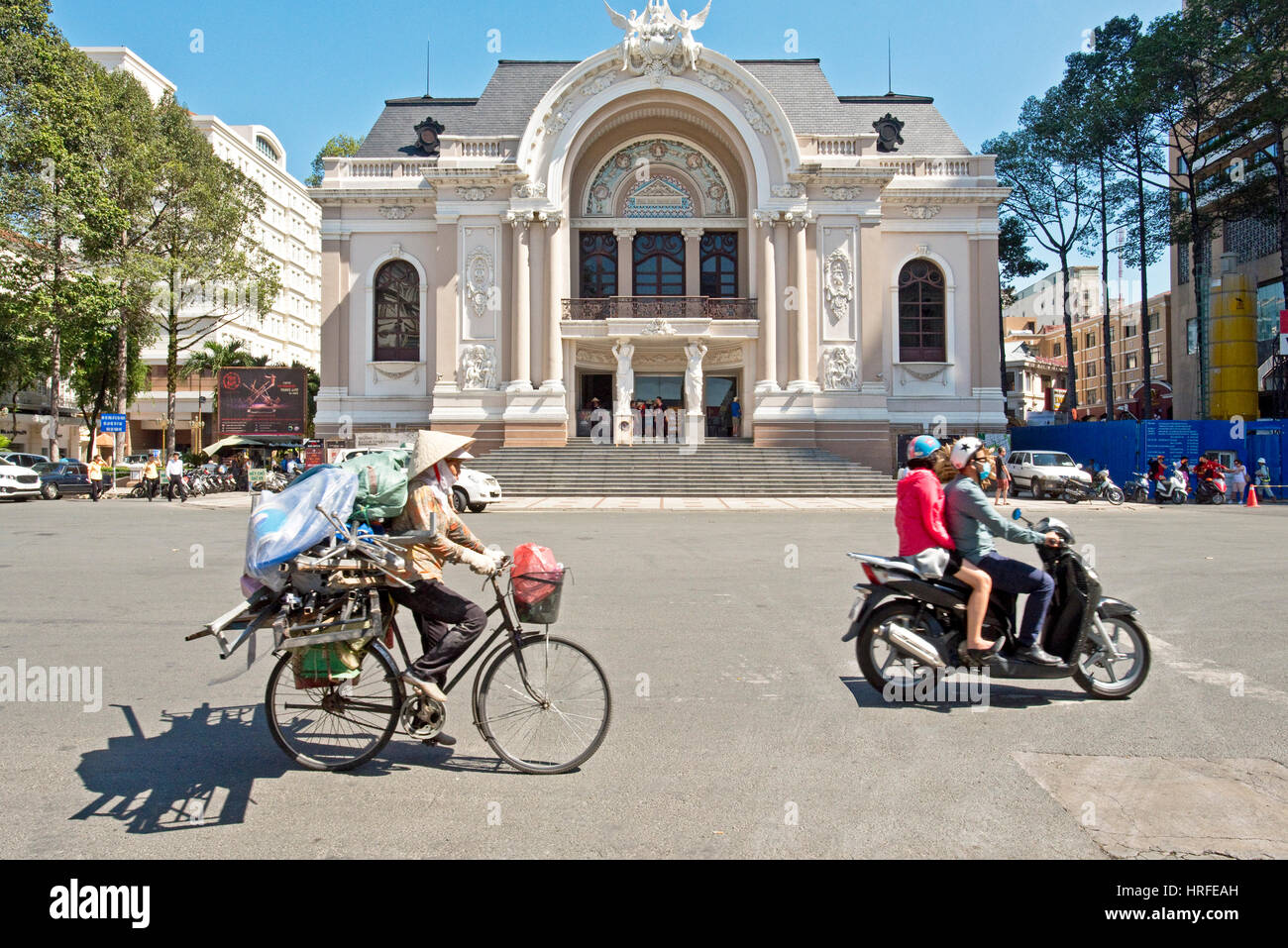 Stadttheater in Ho-Chi-Minh-Stadt mit einheimischen vorbeifahren auf einem Moped und Fahrrad einen traditionellen Hut an einem sonnigen Tag mit blauem Himmel. Stockfoto