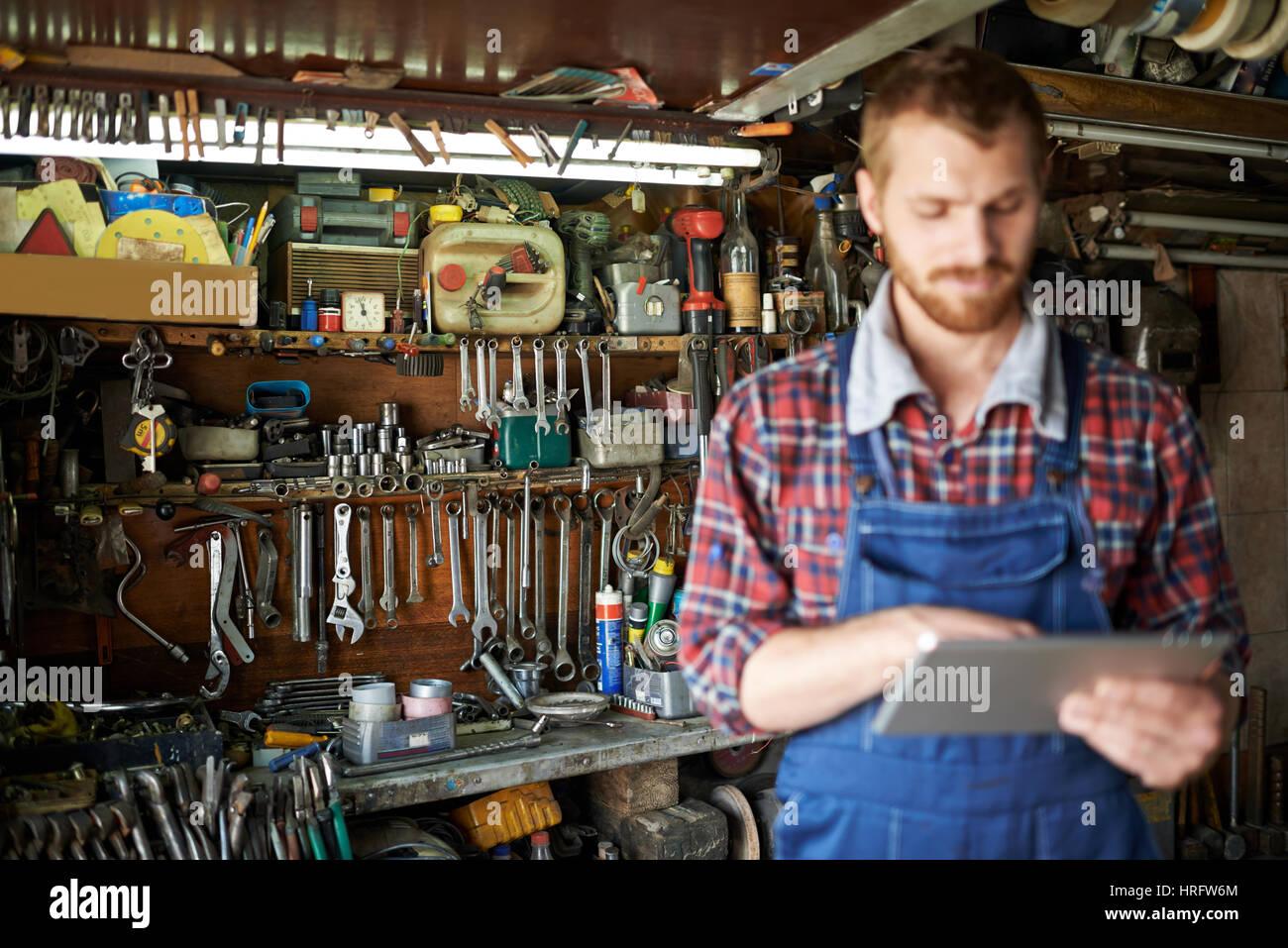 Moderne bärtigen Mechaniker mit Tablet in Werkstatt, Fokus auf Fachböden mit Geräten und Werkzeugen hinter ihm Stockfoto