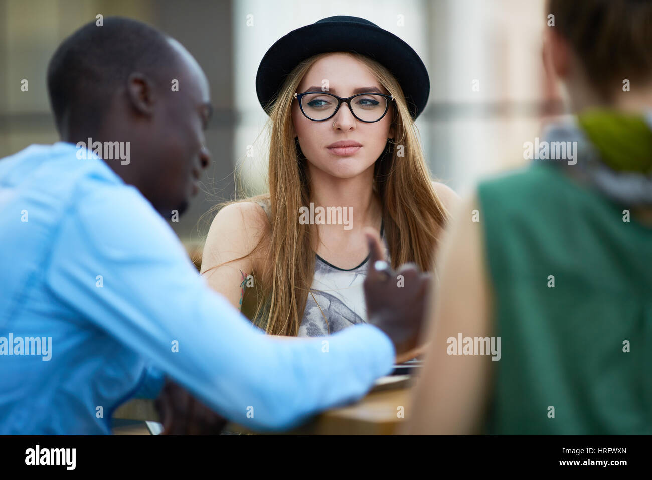 Porträt des jungen stilvolle Mädchen mit schwarzen Hut und Brille sitzt am Tisch im Freien mit ihren Kollegen Stockfoto