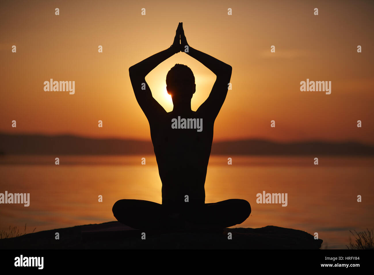 Silhouette der Mann sitzt in Lotus Pose mit seine Arme über Kopf im Namaste und Sonnenuntergang am Wasser Hintergrund gehoben Stockfoto