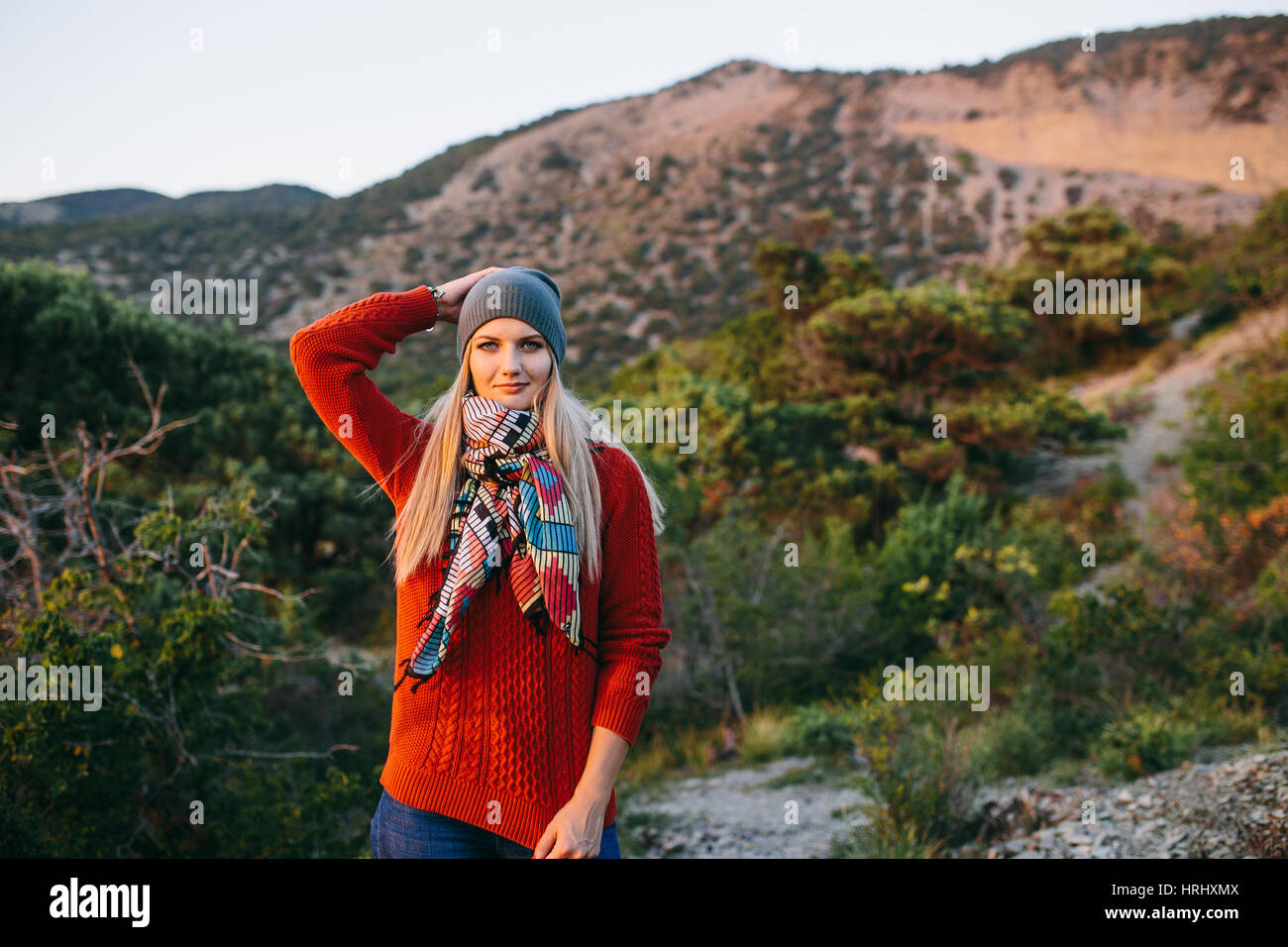 Porträt von eine schöne junge blonde Frau in Mütze, roter Pullover und bunten Schal im Freien auf dem Hintergrund der Berge und Wald Stockfoto