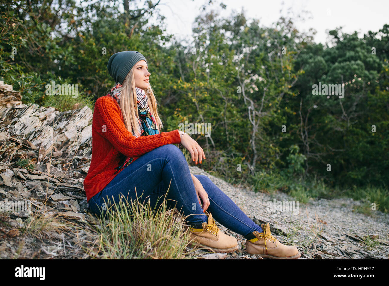 Porträt von eine schöne junge blonde Frau in Hut, roter Pullover, blaue Jeans und gelbe Stiefel sitzt im Freien auf dem Hintergrund des Waldes Stockfoto