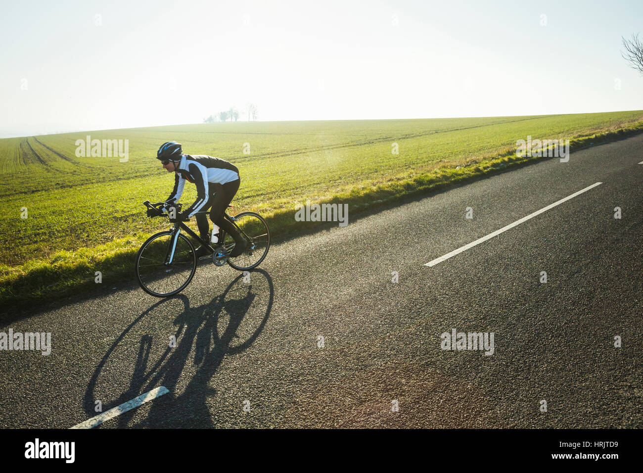 Ein Radsportler entlang einer Landstraße, an einem klaren sonnigen Wintertag. Schatten Sie auf der Fahrbahn. Stockfoto