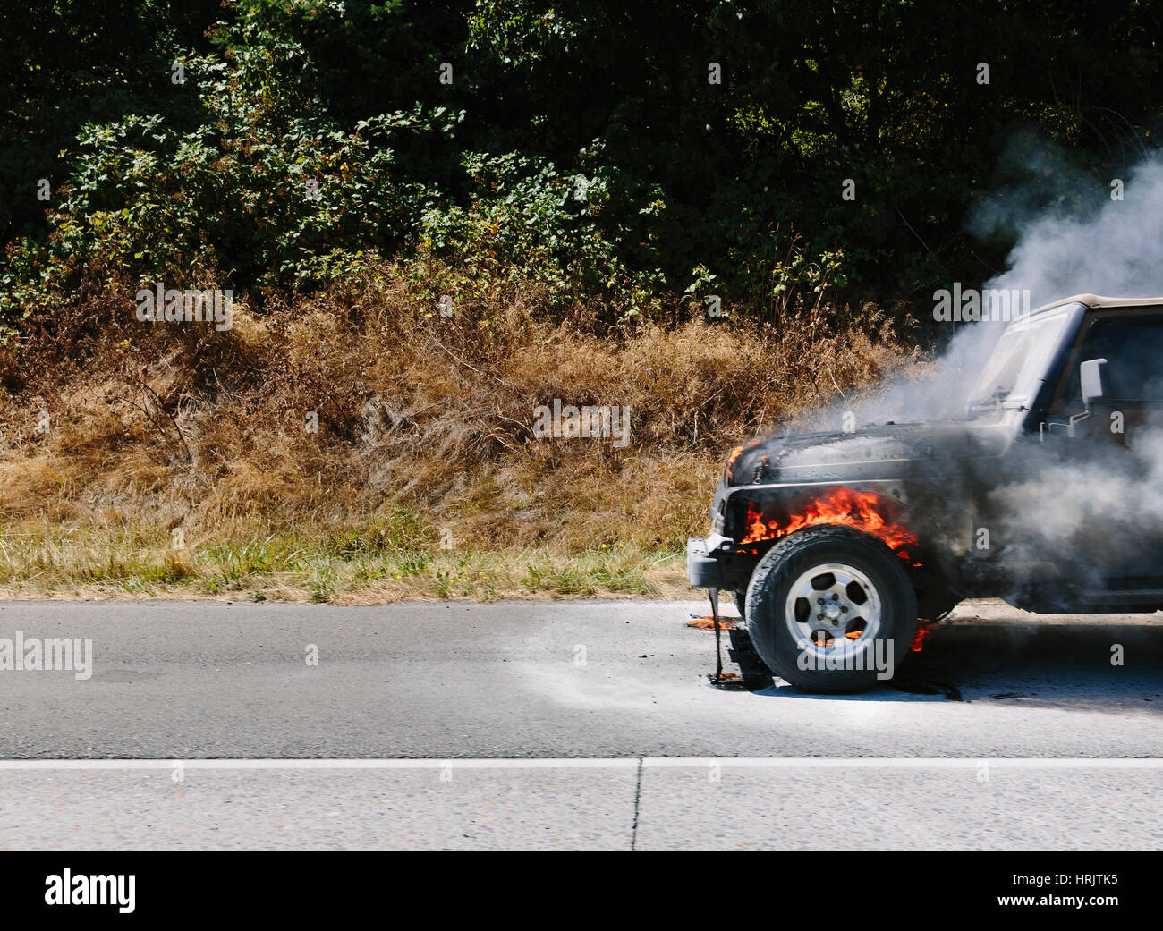 Eine brennende Auto auf einer Landstraße vor Büschen und Bäumen. Stockfoto