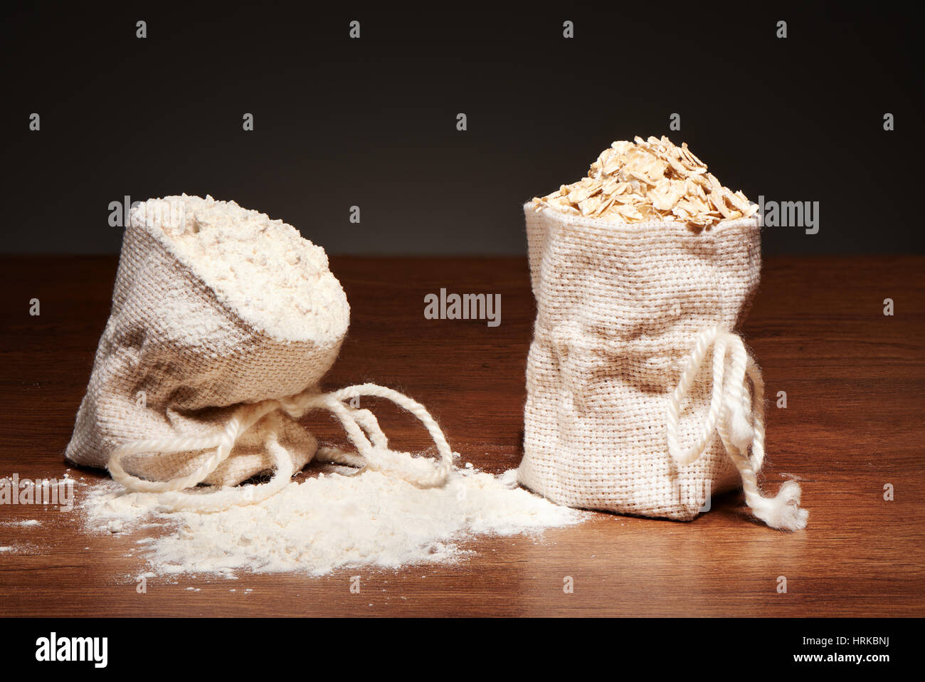 Jute Säcke mit Mehl und Haferflocken Flocken auf dunklen Holztisch auf grauem Hintergrund Stockfoto