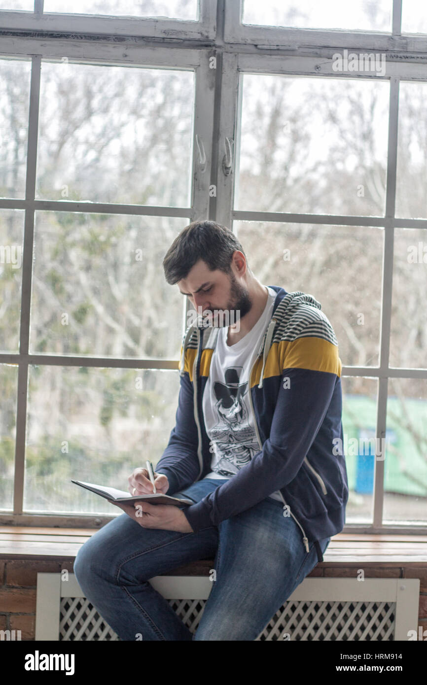 Porträt eines lässig bärtige Hipster schreiben und Notizen im Heft oder Notepad zu Hause. Große Fenster Backdround. Handschrift Stockfoto