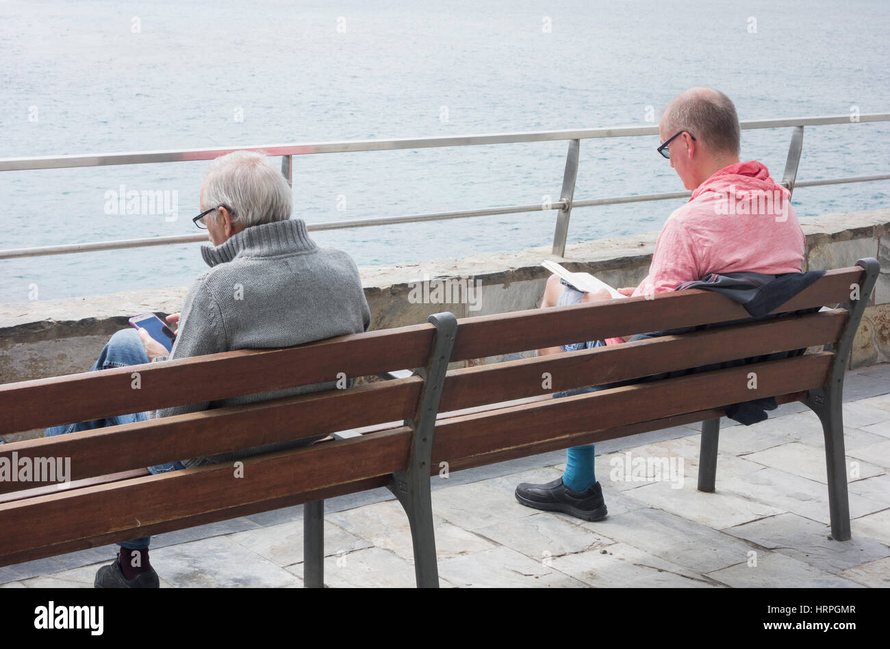 Zwei Männer lesen mit Blick aufs Meer. Ein Taschenbuch und das andere mit mobilen Gerät. Stockfoto