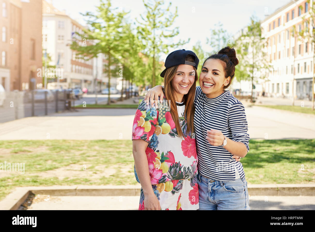 Süße junge Freundinnen posieren Arm in Arm in trendige Sommerkleidung fröhlich grinsend in die Kamera in einer städtischen Straße Stockfoto