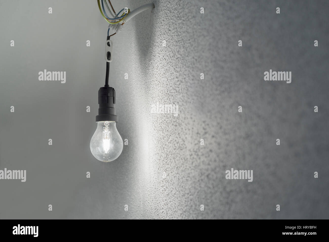 Beleuchtete Halogen Glühbirne in eine schwarze Kunststoff Halter Lampenfassung mit Stromkabeln ragen aus einer Wand mit weißem groben Putz Stockfoto
