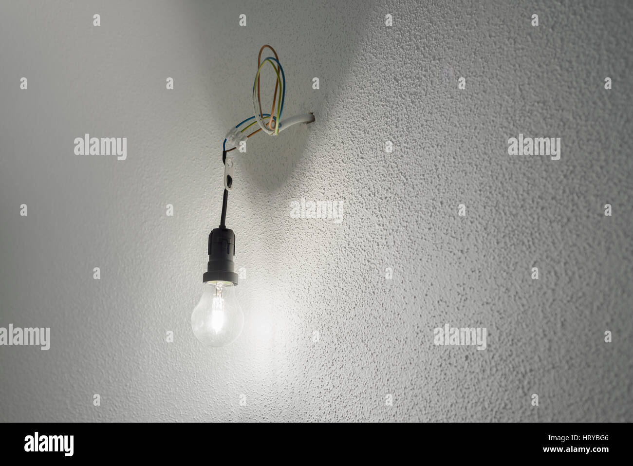 Beleuchtete Halogen Glühbirne in eine schwarze Kunststoff Halter Lampenfassung mit Stromkabeln ragen aus einer Wand mit weißem groben Putz Stockfoto