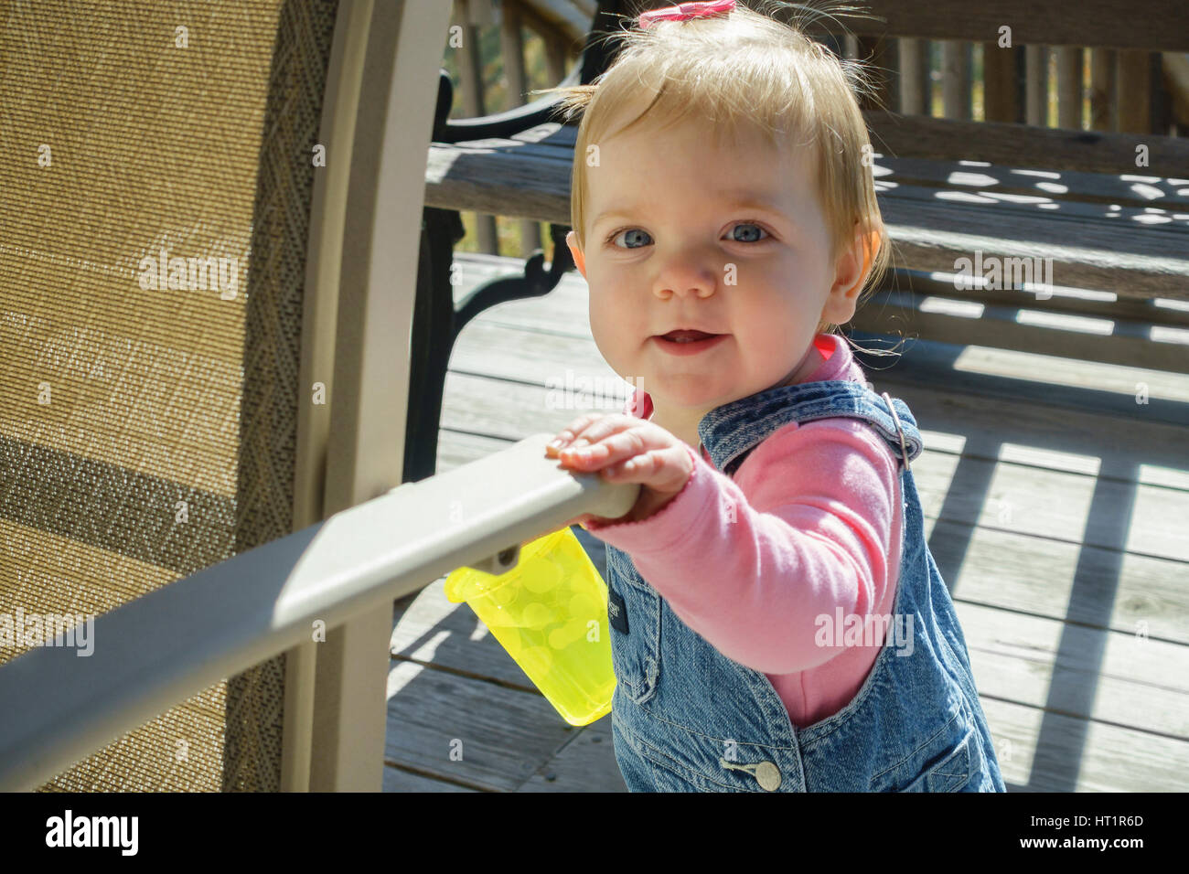 Ein Lächeln auf den Lippen kaukasischen Baby Einjahresmädchen im Overall mit einem Getränk, stehend auf einer Außenterrasse mit Sonnenschein. Kansas, USA. Stockfoto