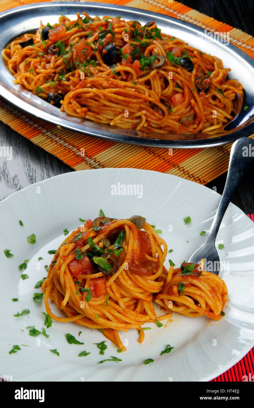Spaghetti Puttanesca auf bunte Tischsets liegen auf Holztisch - italienisches Rezept mit Knoblauch, Olivenöl, Sardellen, Oliven, Tomaten, Petersilie, Kapern Stockfoto