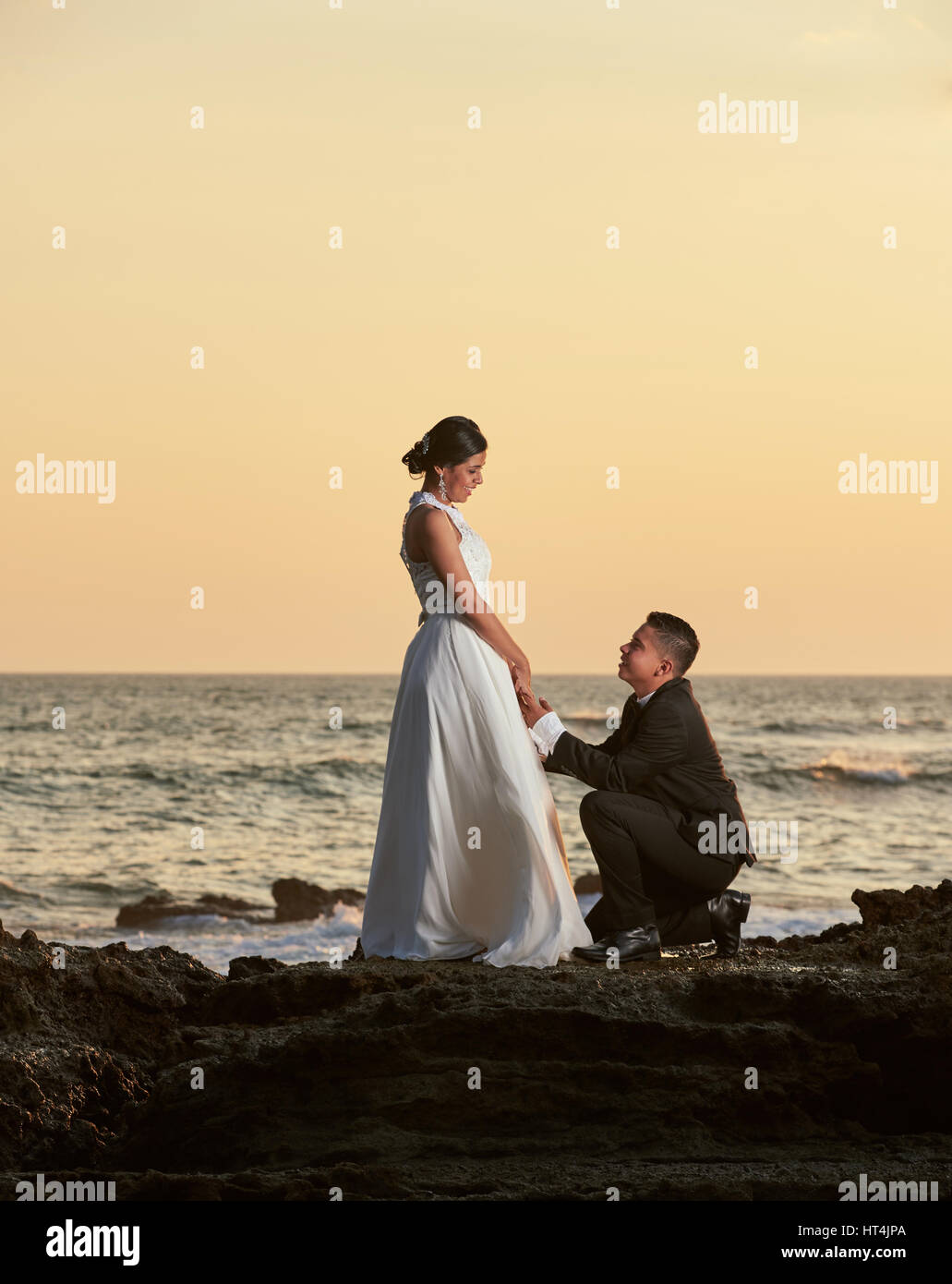 Bräutigam auf Knien stehen und halten Braut Hand auf orange Sonnenuntergang Hintergrund im Meeresstrand Stockfoto