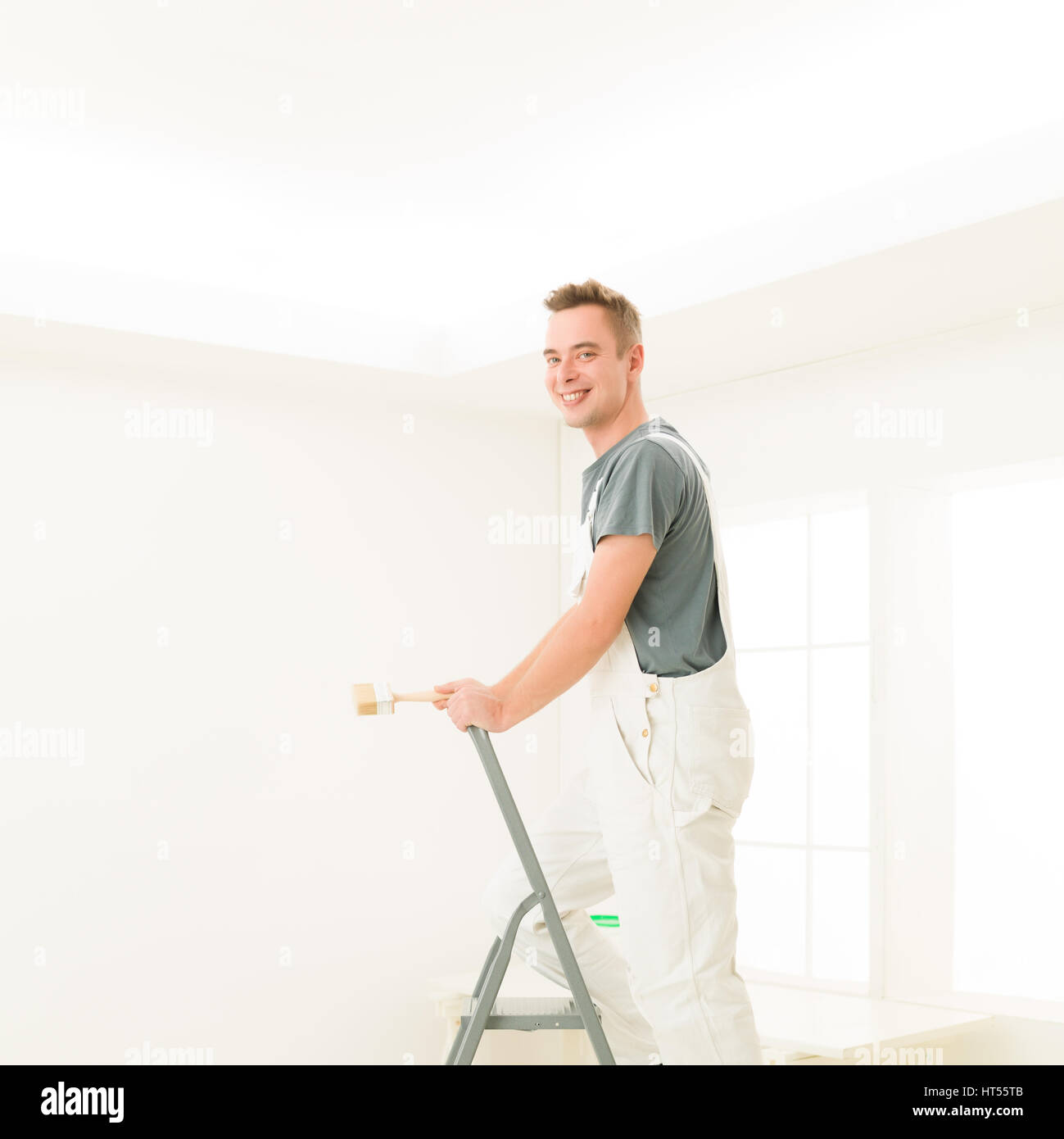 Quadratische Porträt junger Mann Lachen stehen auf einer Leiter mit einem Pinsel, strahlend weiß zu Hause. Reparatur, Bau, Renovierung und Menschen-Konzept Stockfoto