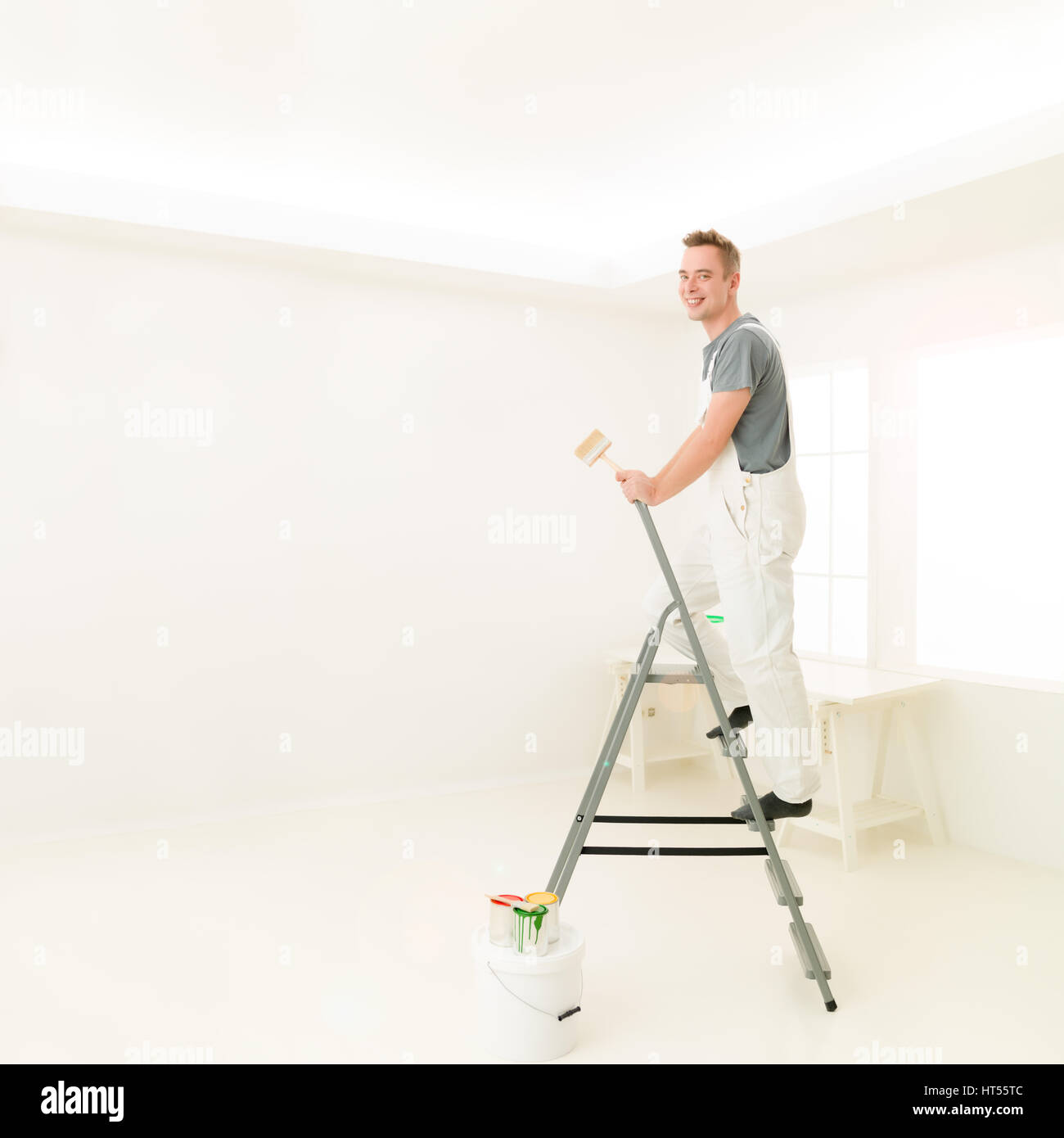 Quadratische Porträt junger Mann Lachen stehen auf einer Leiter mit einem Pinsel, strahlend weiß zu Hause. Reparatur, Bau, Renovierung und Menschen-Konzept Stockfoto