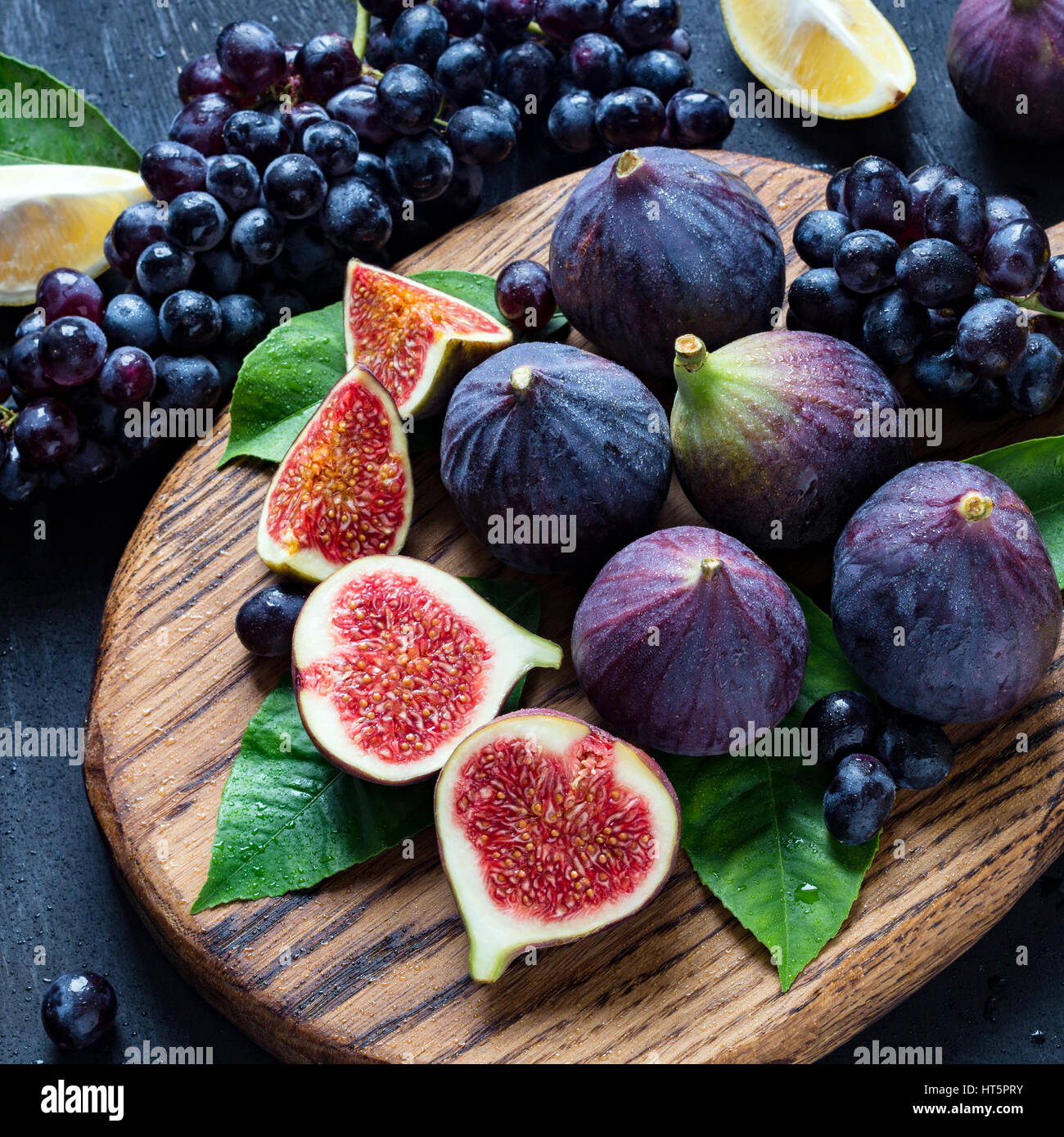 Frische schwarze Trauben, Zitronen und lila Feigen auf Holzbrett mit Wasser besprüht. Stillleben mit frischen Früchten. Quadratische Ernte Stockfoto