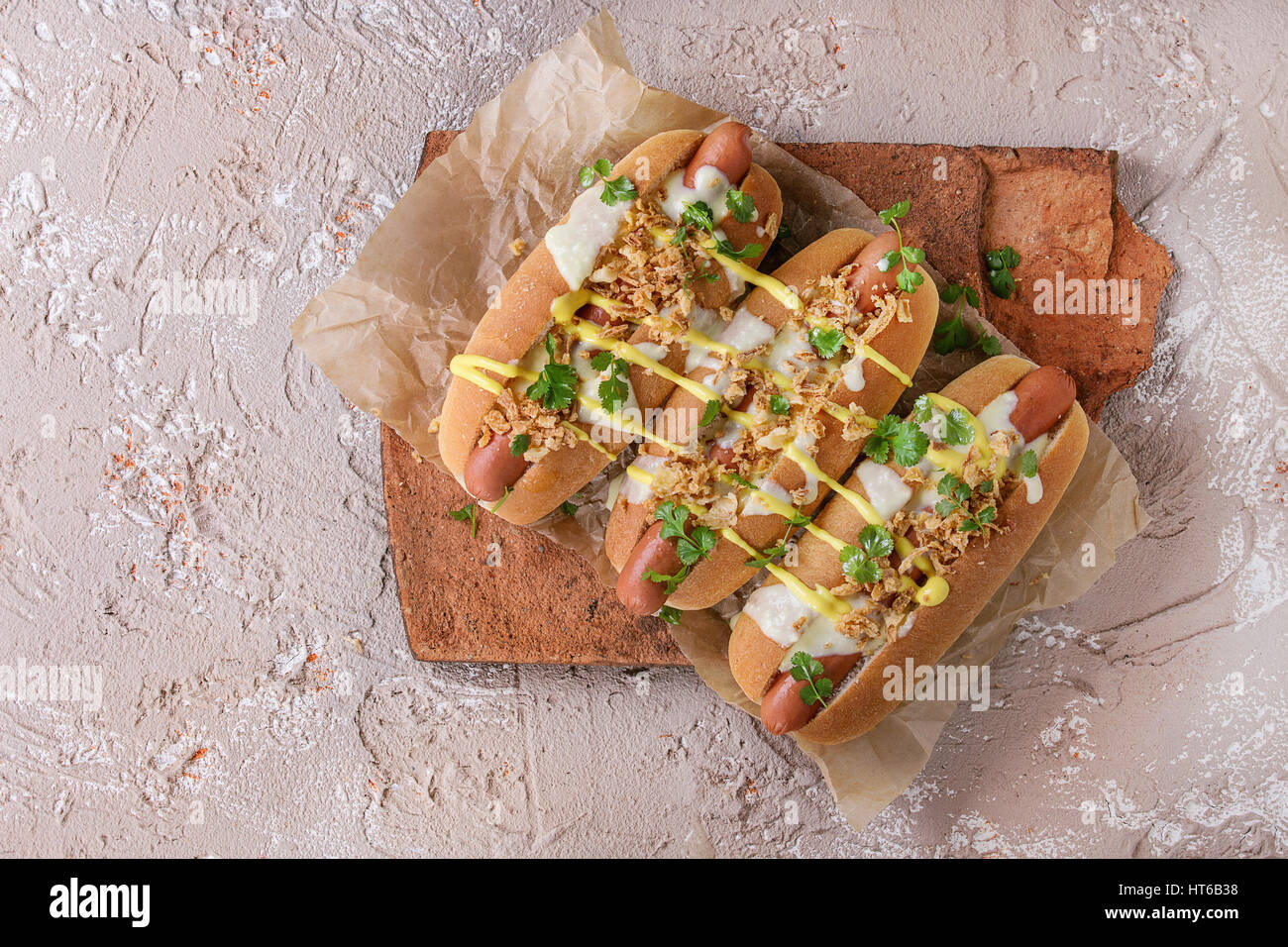 Hot Dogs mit Wurst, gebratenen Zwiebeln, Koriander, Käse-Sauce und Senf, serviert auf Terrakotta-Board über Beige Betontextur Hintergrund. FAS Stockfoto