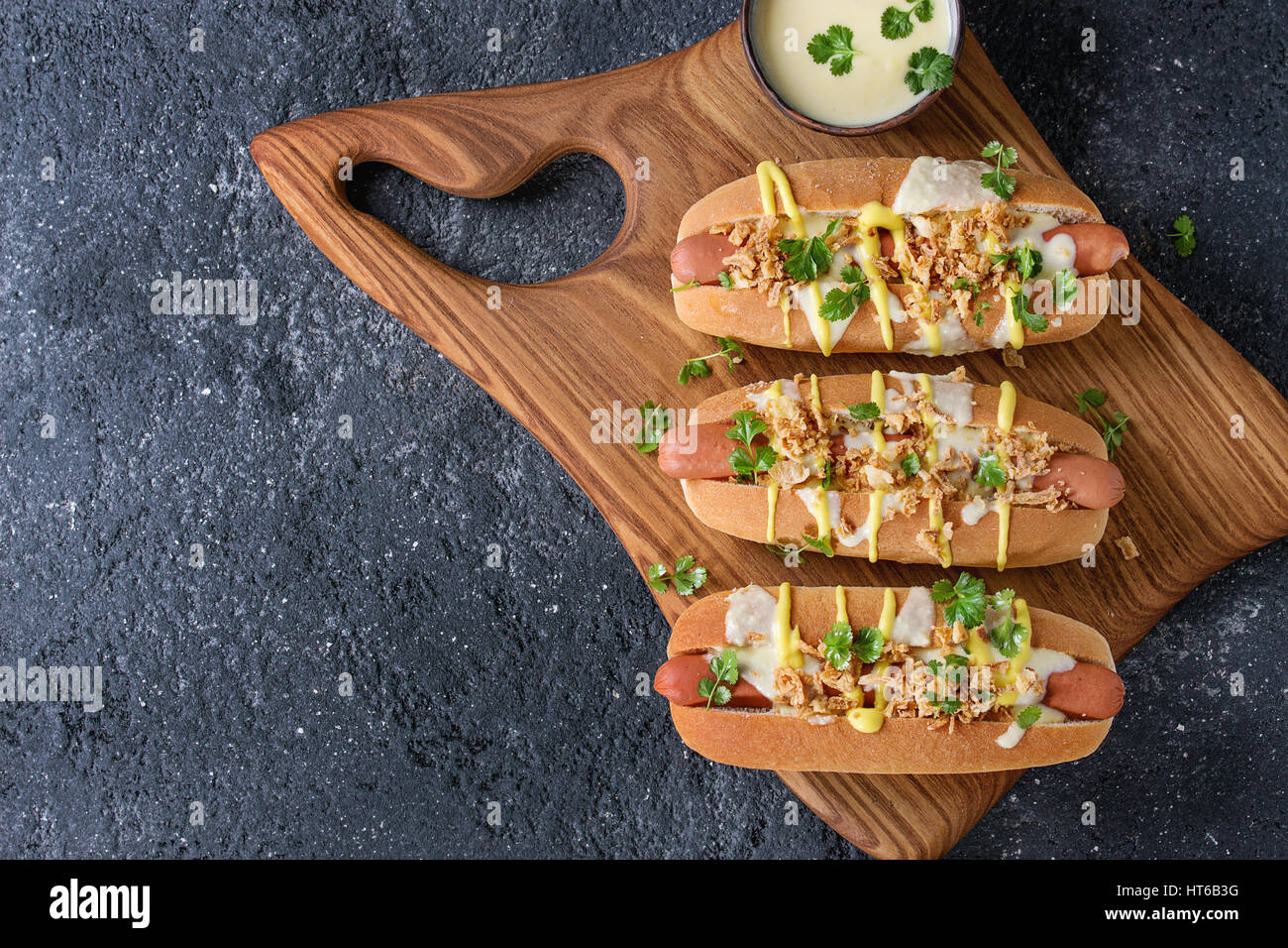 Hot Dogs mit Wurst, gebratenen Zwiebeln, Koriander, Käse-Sauce und Senf, serviert auf hölzernen Portion Brett über schwarze Betontextur Hintergrund. Stockfoto