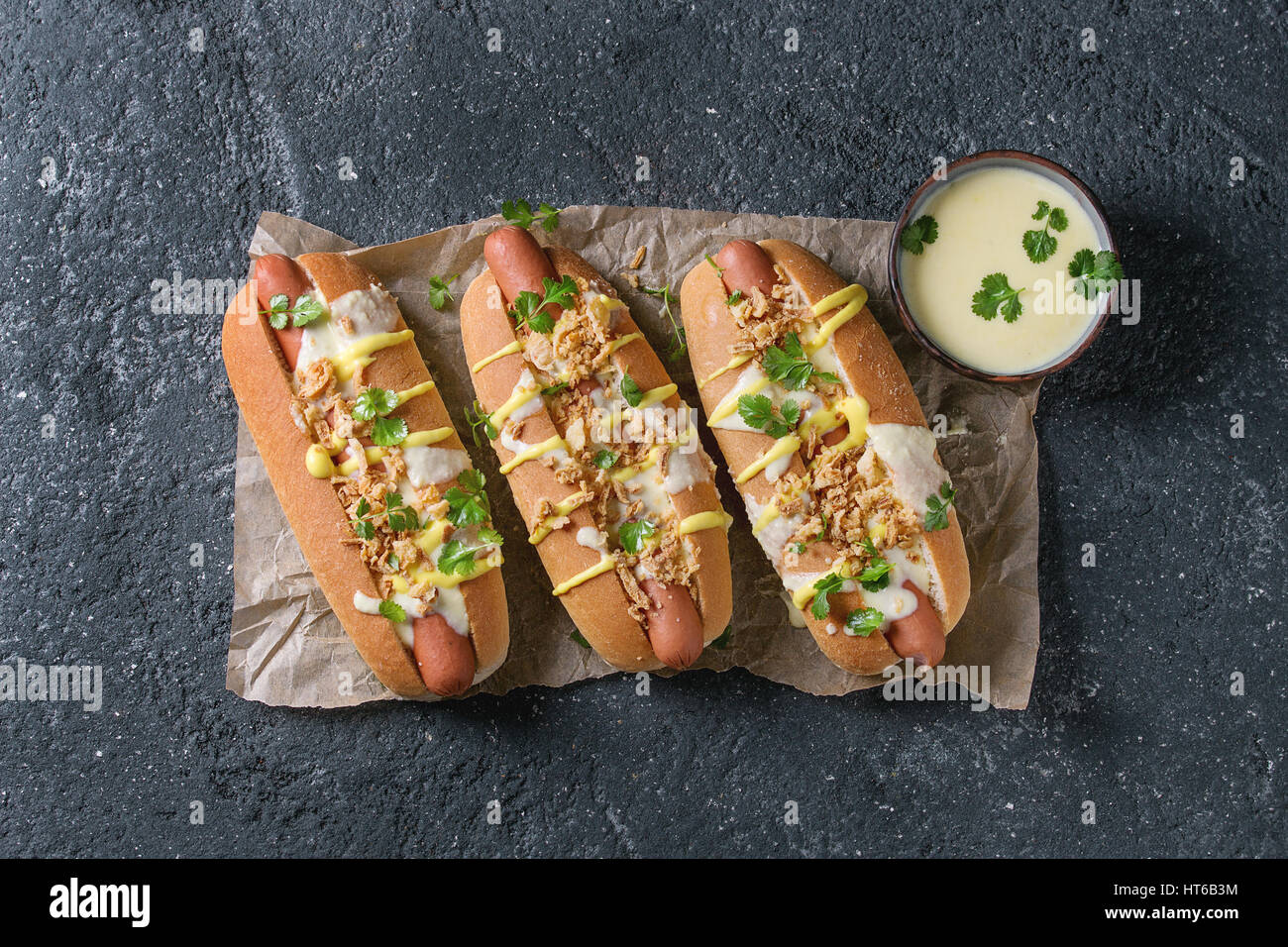 Hot Dogs mit Wurst, gebratenen Zwiebeln, Koriander, Käse-Sauce und Senf, serviert auf Backpapier über schwarze Betontextur Hintergrund. Schnell fo Stockfoto