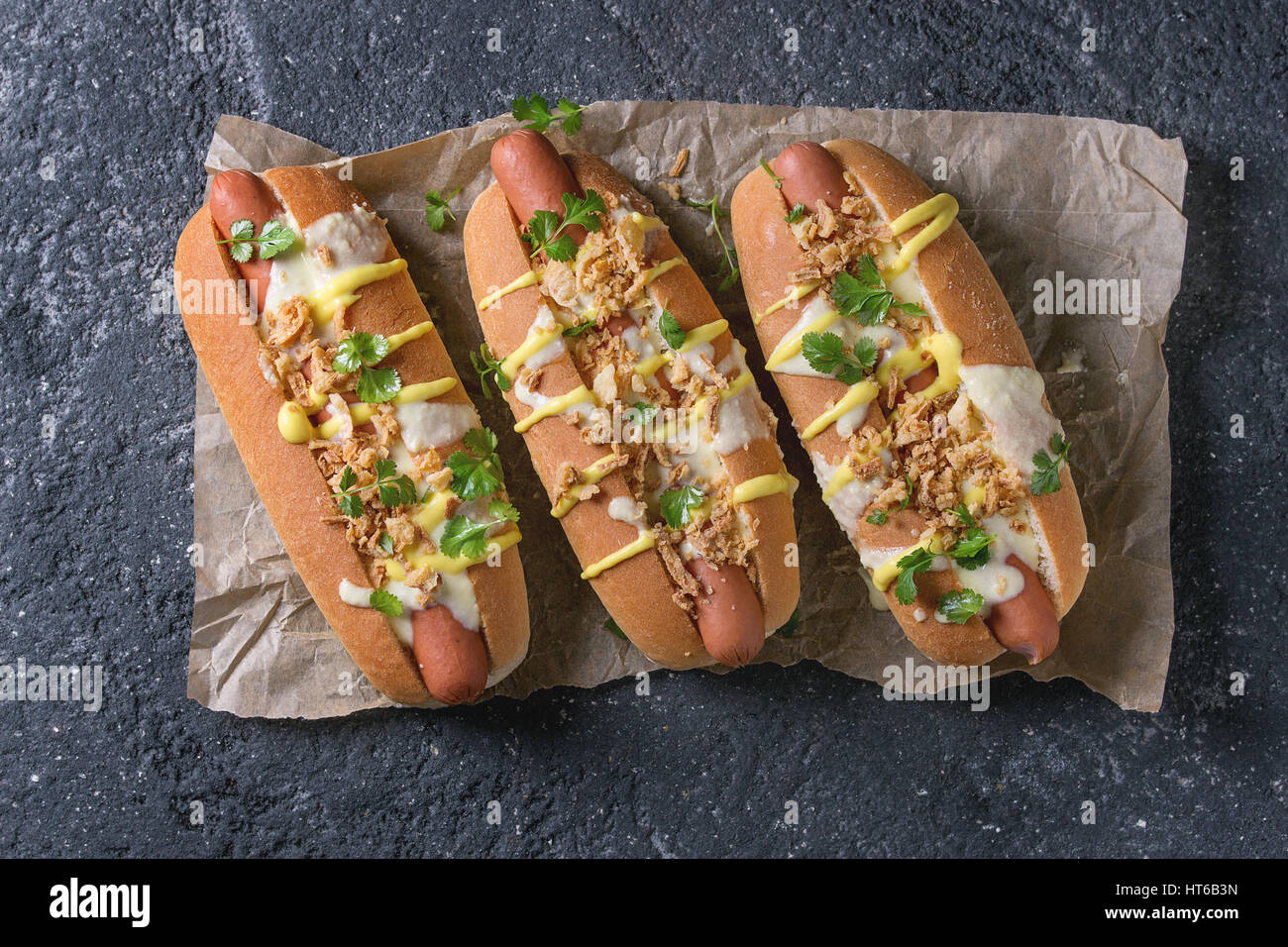 Hot Dogs mit Wurst, gebratenen Zwiebeln, Koriander, Käse-Sauce und Senf, serviert auf Backpapier über schwarze Betontextur Hintergrund. Schnell fo Stockfoto