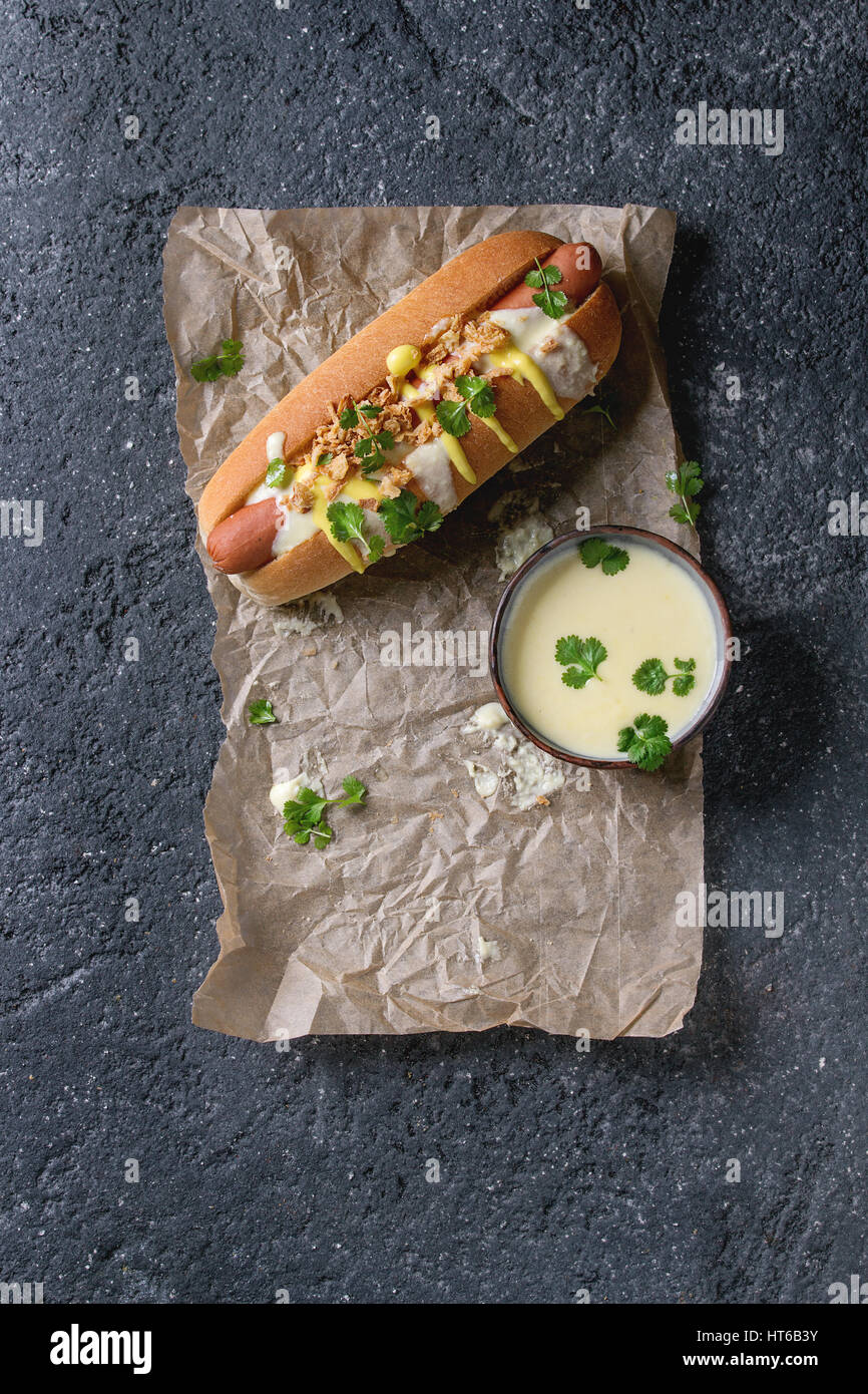 Ein Hot Dog mit Wurst, gebratenen Zwiebeln, Koriander, Käse-Sauce und Senf, serviert auf Backpapier über schwarze Betontextur Hintergrund. Schnell Stockfoto