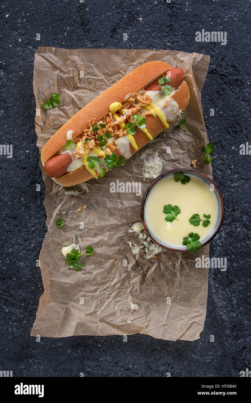 Ein Hot Dog mit Wurst, gebratenen Zwiebeln, Koriander, Käse-Sauce und Senf, serviert auf Backpapier über schwarze Betontextur Hintergrund. Schnell Stockfoto