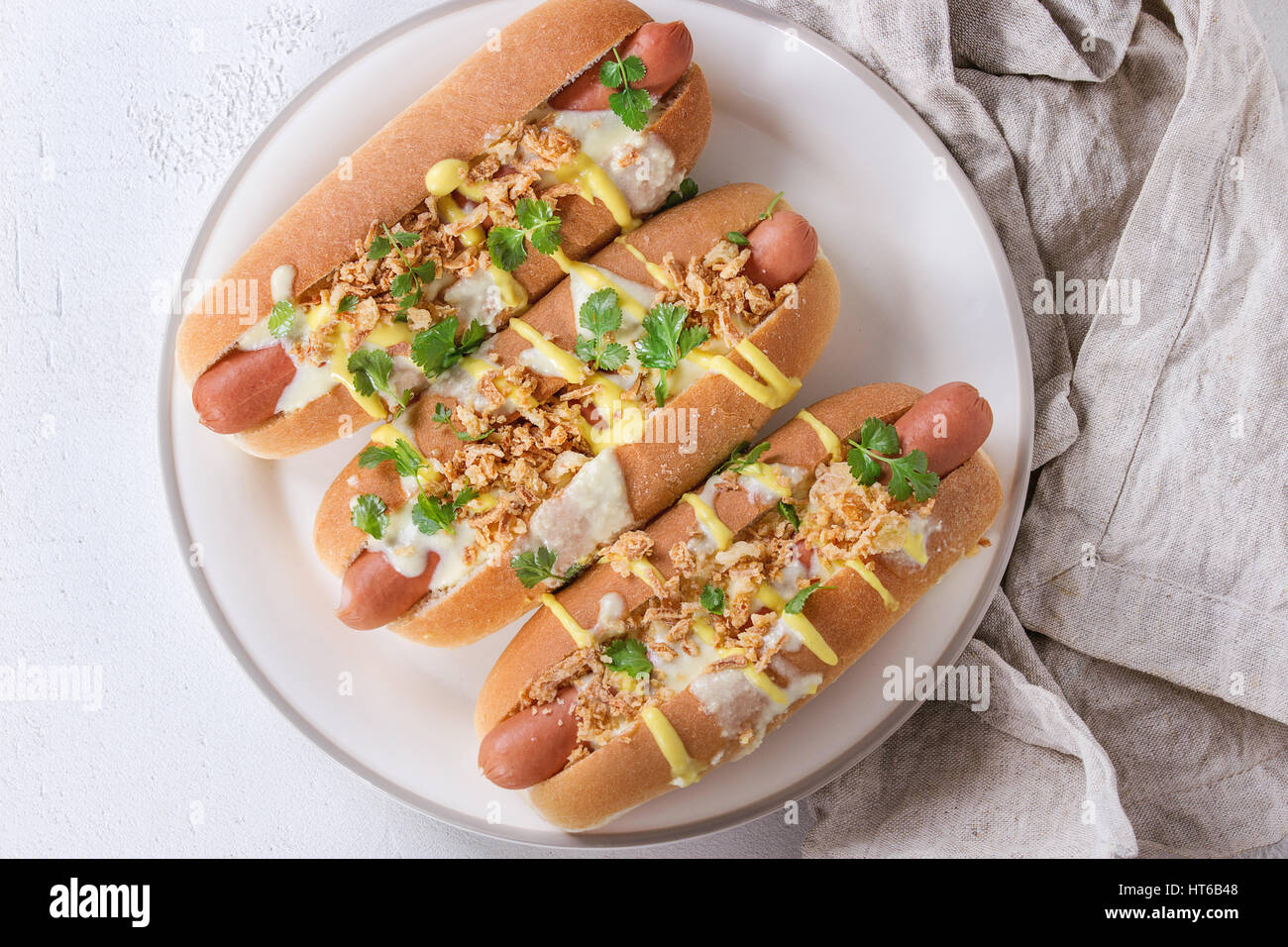 Hot Dogs mit Wurst, gebratenen Zwiebeln, Koriander, Käse-Sauce und Senf, serviert auf weißen Keramikplatte mit Textil über weißem Beton Textur Stockfoto