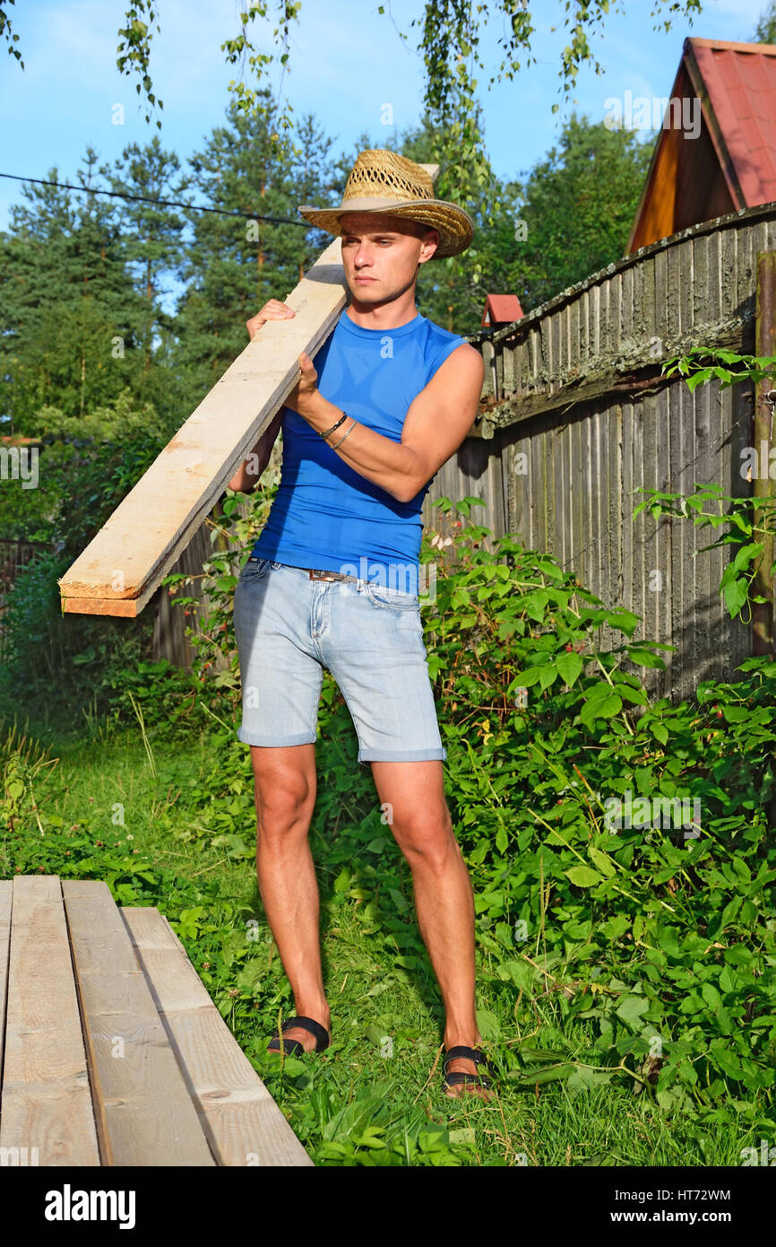 Ein junger Mann mit der Kiefer Board auf seiner Schulter über Berge von Brettern auf dem Grundstück im Sommer an einem sonnigen Tag. Stockfoto