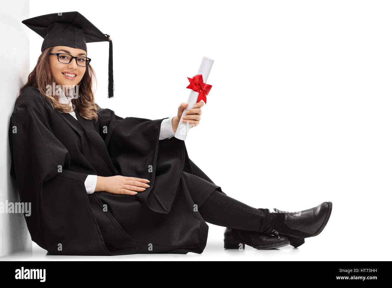 Glückliche Studentin mit einem Diplom auf dem Boden sitzend und lehnte sich gegen eine Wand isoliert auf weißem Hintergrund Stockfoto