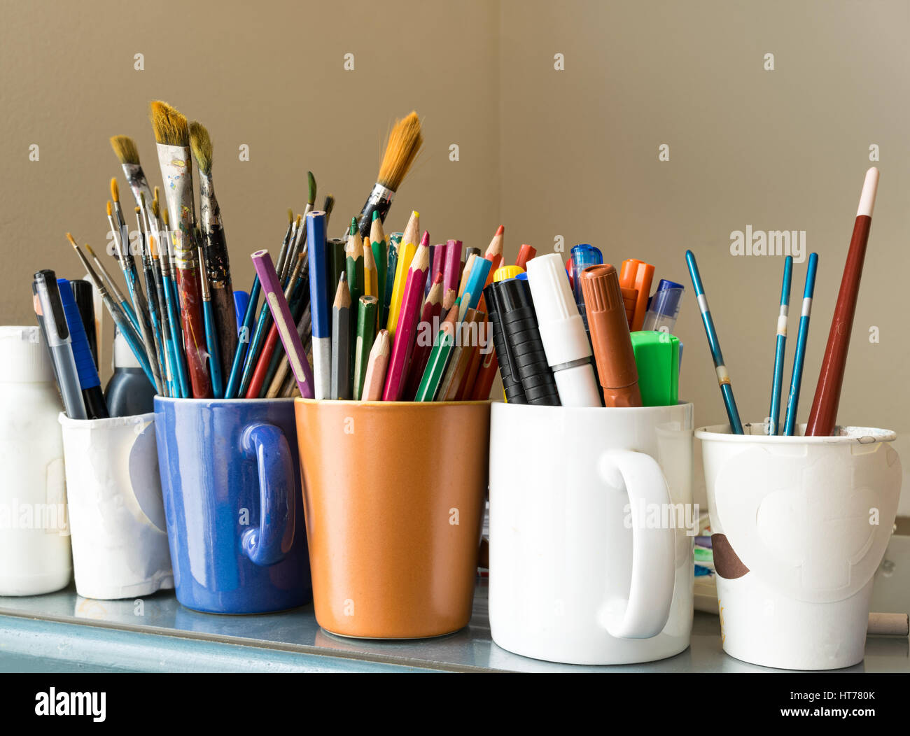 Nahaufnahme der verschiedenen verwendeten Pinsel, geschärften Buntstifte, Kugelschreiber und Marker auf farbige Tassen über blauer Tisch und Beige Hintergrund Stockfoto