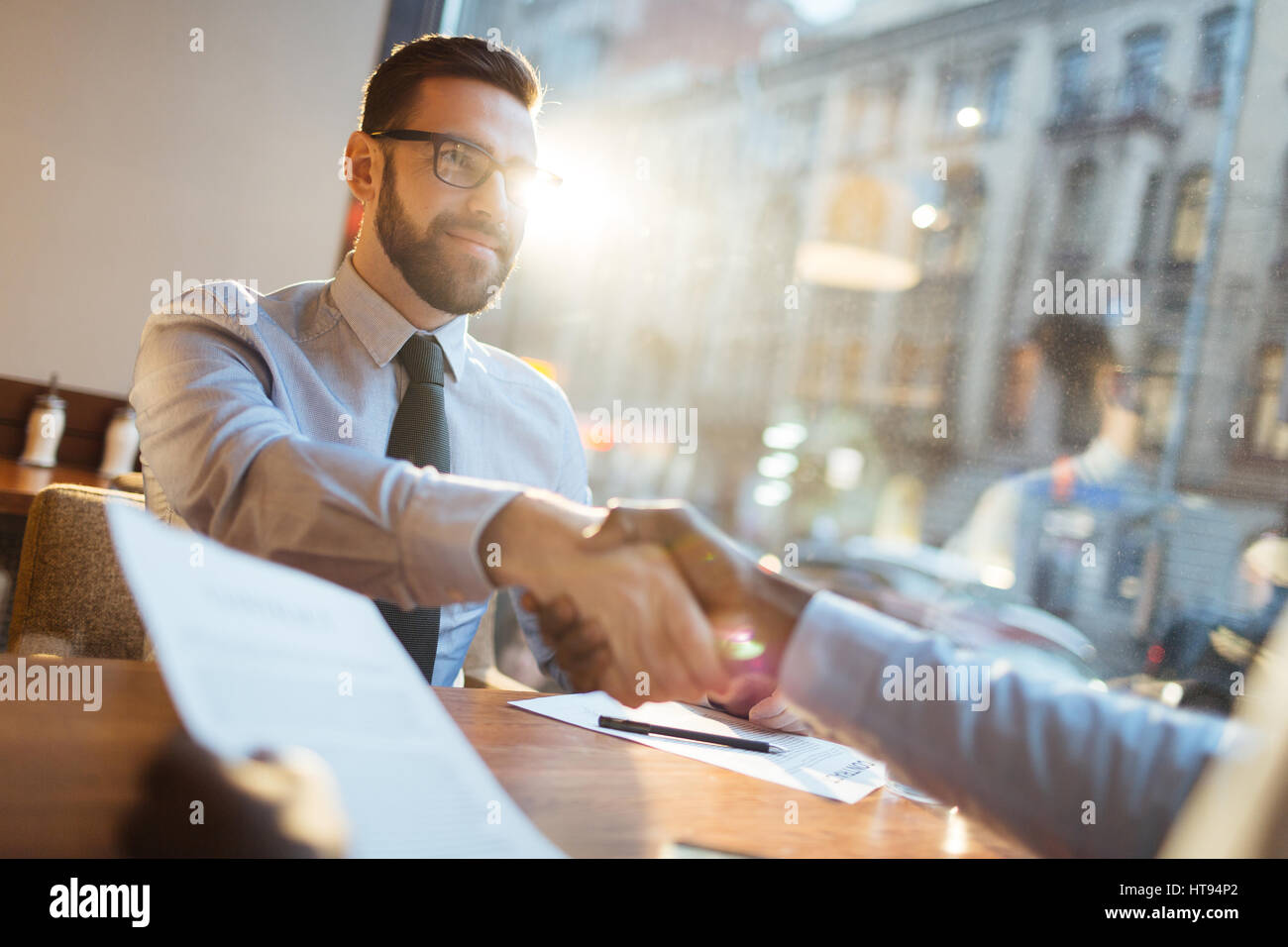 Zuversichtlich bärtigen Geschäftsmann in Brillen am Café-Tisch sitzen und schüttelte die Hand seines Partners und lächelnd nach profitablen Vereinbarung. Stockfoto