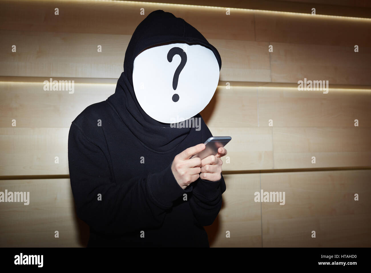 Hacker in schwarzer Hoodie und Fragezeichen Maske mit Smartphone zum Datendiebstahl stehend gegen Holzwand Stockfoto