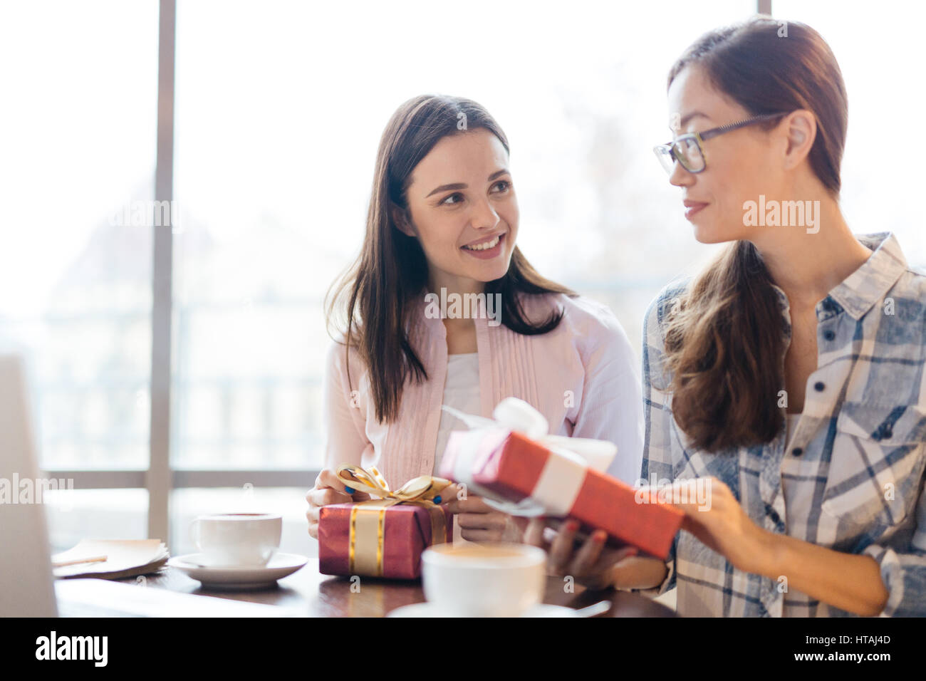 Aufrichtige Szene zweier junger Frauen, eine asiatische, Austausch von Geschenken und Gespräch am Tisch im Café sitzen gegen große Fenster sowohl lächelnd und liebevoll suchen Stockfoto