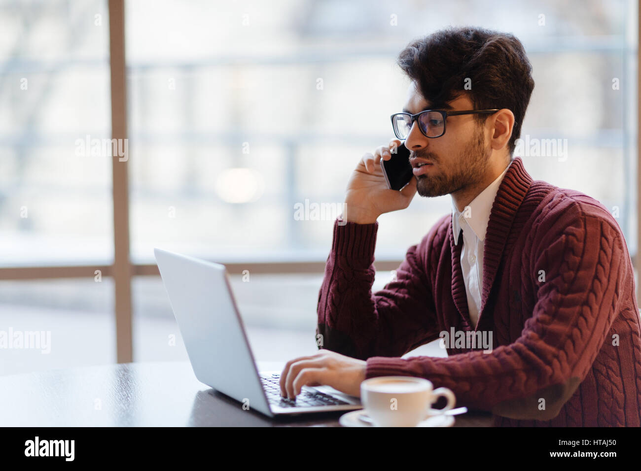 Blick Profilbildnis jungen arabischen Geschäftsmann sitzt am Tisch gegen große Fenster, die Beratung mit seinem Partner über Geschäft per Telefon und ch Stockfoto