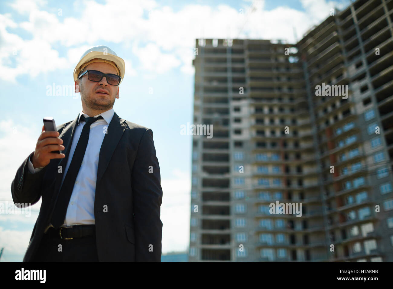 Porträt von erfolgreicher zuversichtlich Geschäftsmann mit harten Hut und Sonnenbrille mit schwarzen Anzug stand Wegschauen gegen Hochhauswohnung Bui Stockfoto