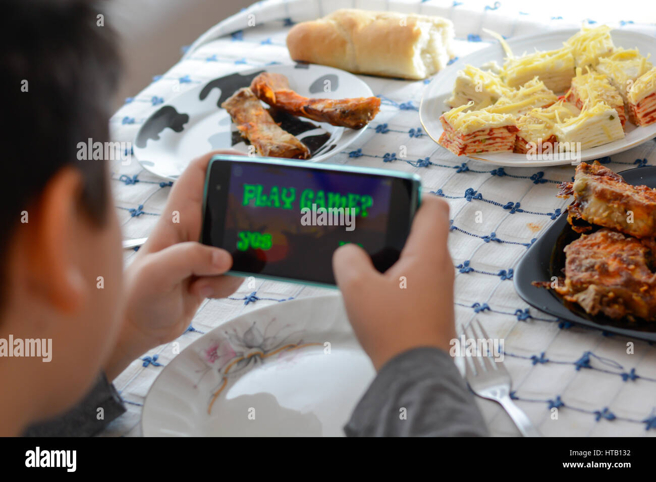 Junge spielt mit seinem Handy in der Mittagspause. Platten mit verschiedenen Lebensmitteln im Hintergrund Stockfoto