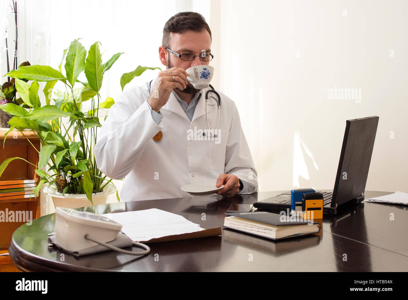 Arzt im Büro sitzen an einem Schreibtisch und trinken Sie eine Tasse Kaffee.  Der Arzt während einer Pause, trinken eine Tasse Kaffee. Stockfoto