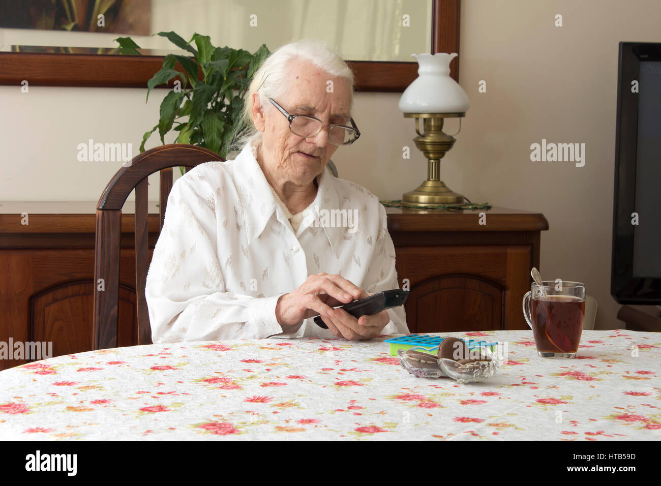 Alte Frau am Tisch mit Fernbedienung für den Fernseher.  Die alte Frau stellt sich auf die TV-Fernbedienung, die am Tisch sitzen.  Die alte Frau in Gläser s Stockfoto