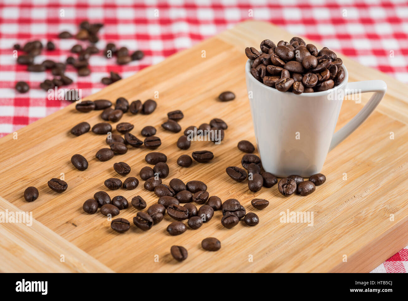 Eine weiße Kaffeetasse mit Kaffeebohnen auf Holz und eine rot-weiß karierte Tischdecke im Hintergrund. Stockfoto