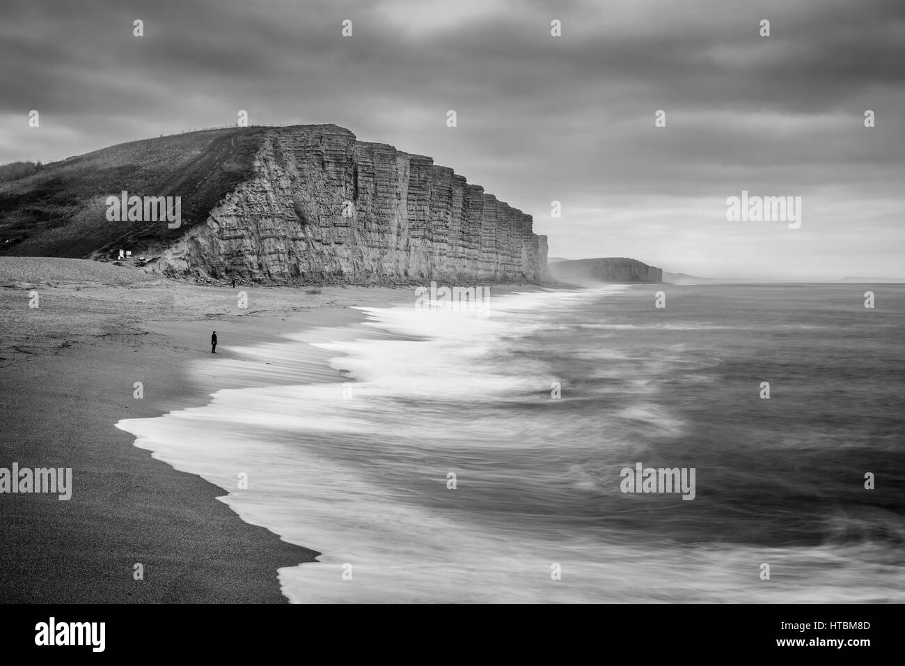 eine einsame Figur gerade Wellen brechen sich am Strand unter East Cliff, West Bay, Jurassic Coast, Dorset, England, UK Stockfoto