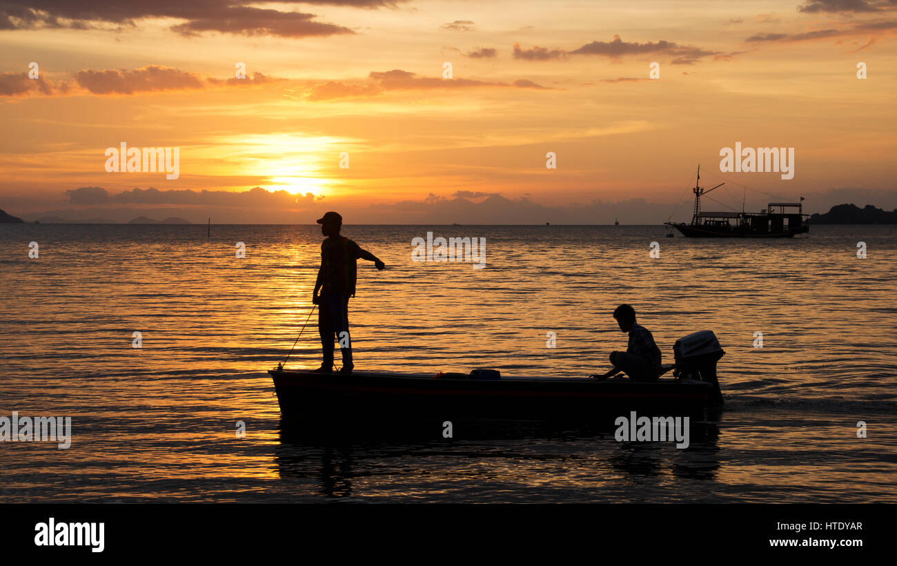 Silhouette von zwei Fischern auf einem kleinen Fischerboot mit Außenbordmotor auf dem Ozean bei Einbruch der Dunkelheit mit einem orange Sonnenuntergang im Hintergrund in Asien. Stockfoto