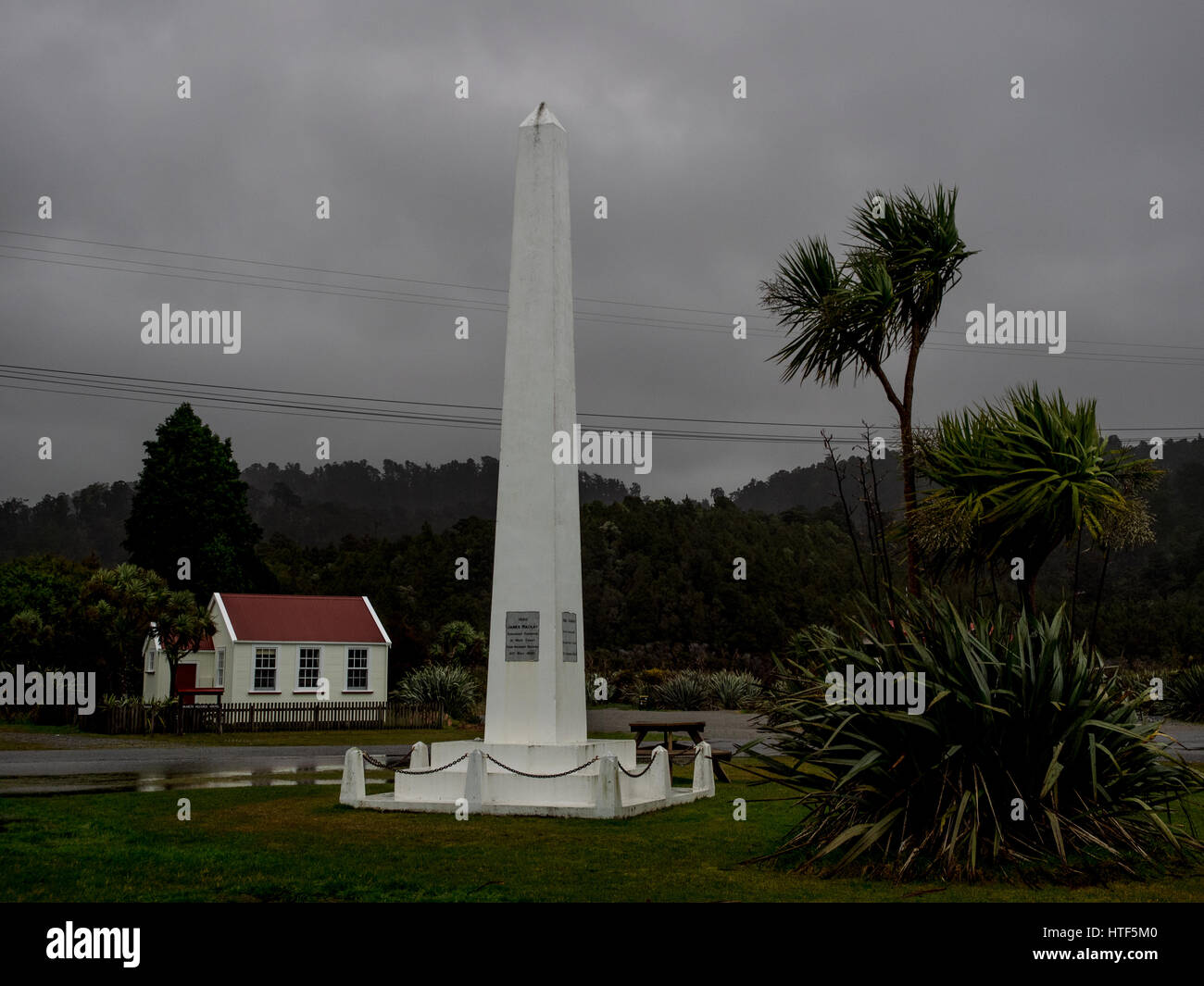 Tasman Denkmal mit historischen Schulhaus hinter Okarito, South Westland, Neuseeland. Einen düsteren Abend Kohl Baum Ti Kouka verlässt Schlag im Wind. Stockfoto