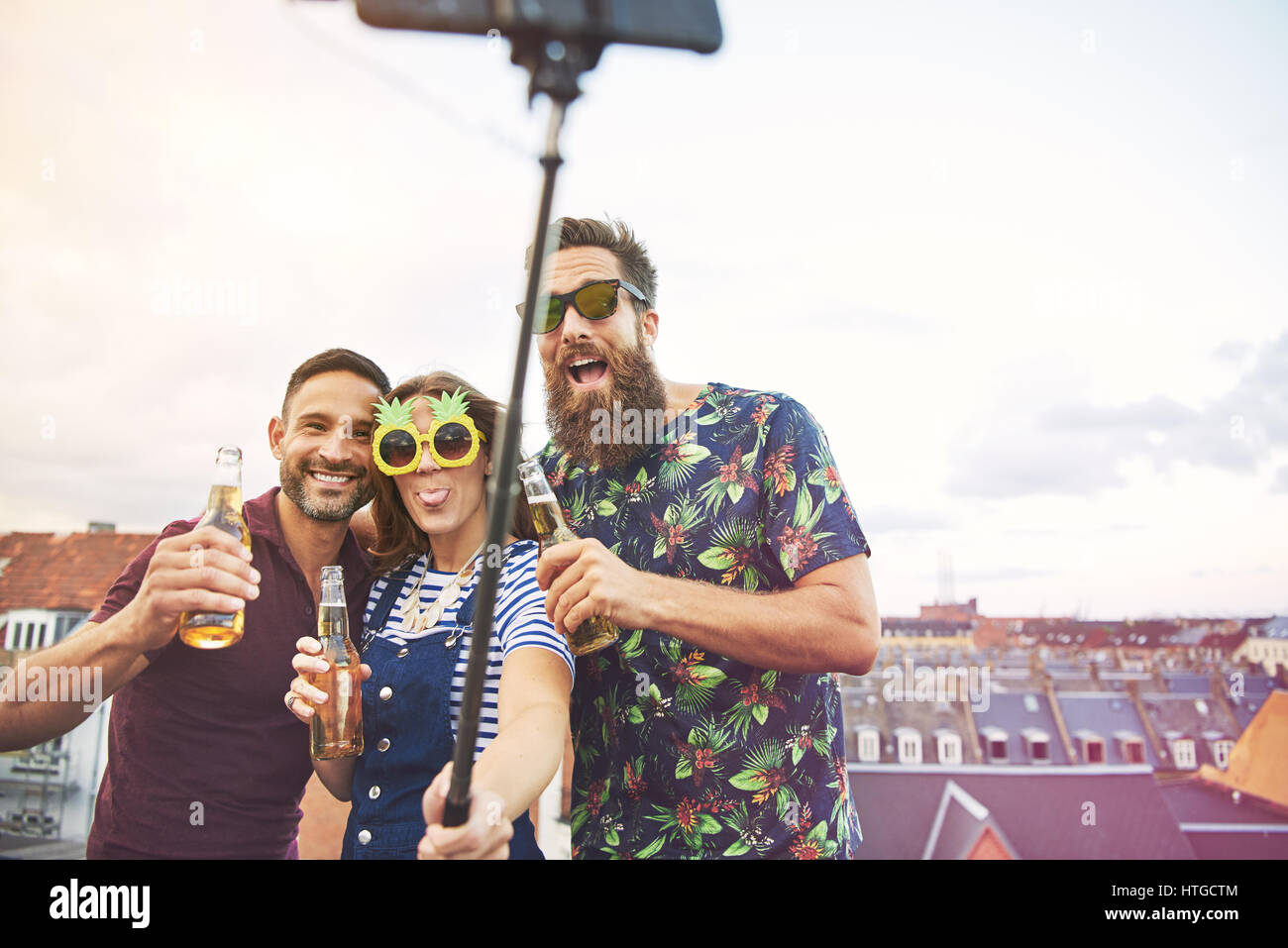 Gruppe von drei betrunkene Jugendliche trinken und nehmen Bilder von sich selbst auf Dach mit Selfie Stick auf Dach mit textfreiraum in hellen Himmel Stockfoto