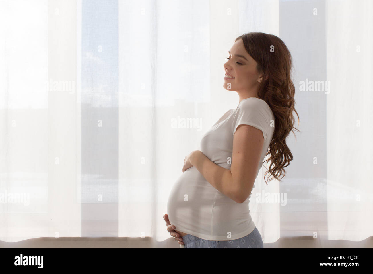 Junge schwangere Frau mit geschlossenen Augen ihren Magen zu umarmen. Horizontale drinnen geschossen Stockfoto